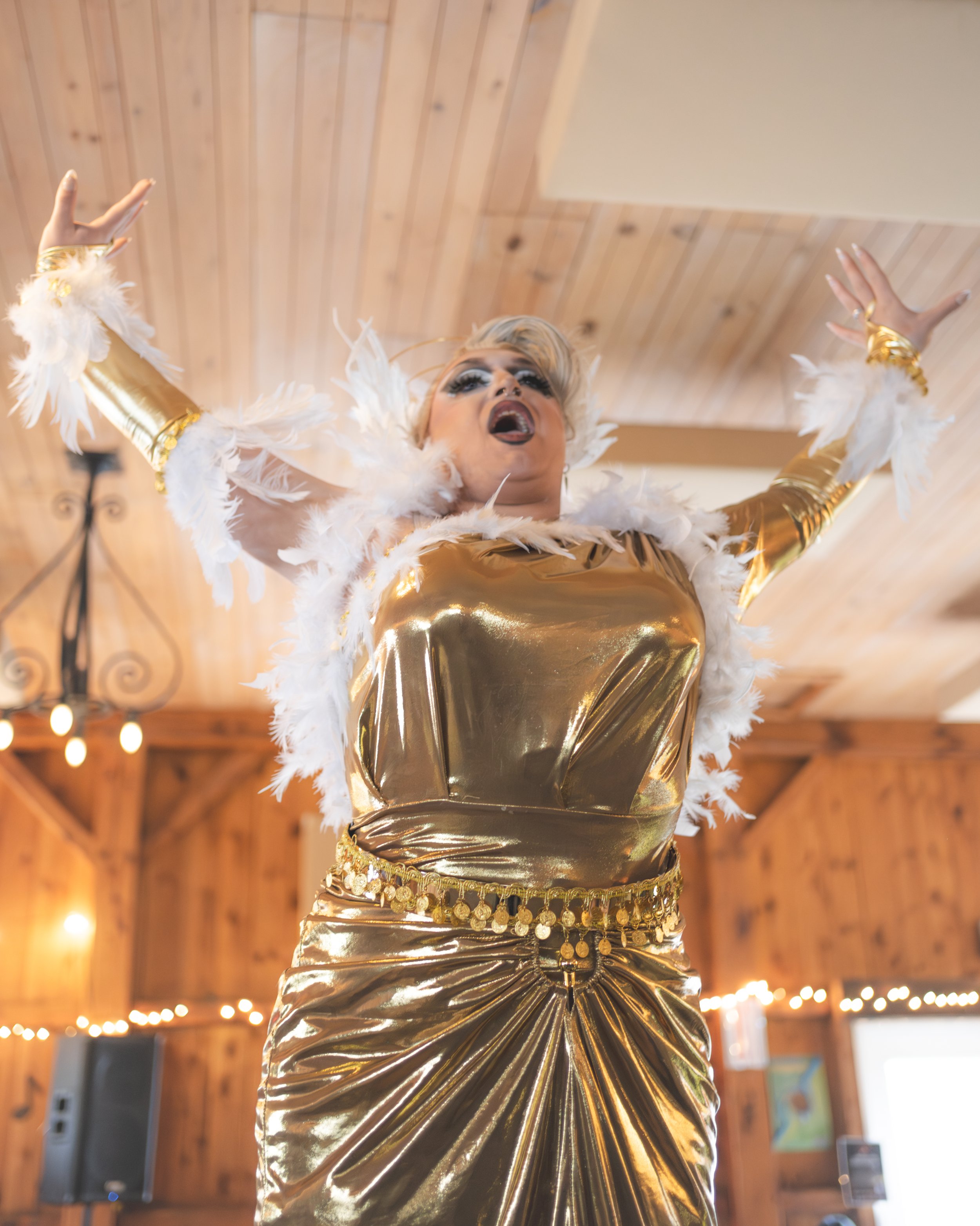 Performer dressed in a shiny gold dress with feathered details, raising her arms with expressive gestures in a wooden-paneled room.
