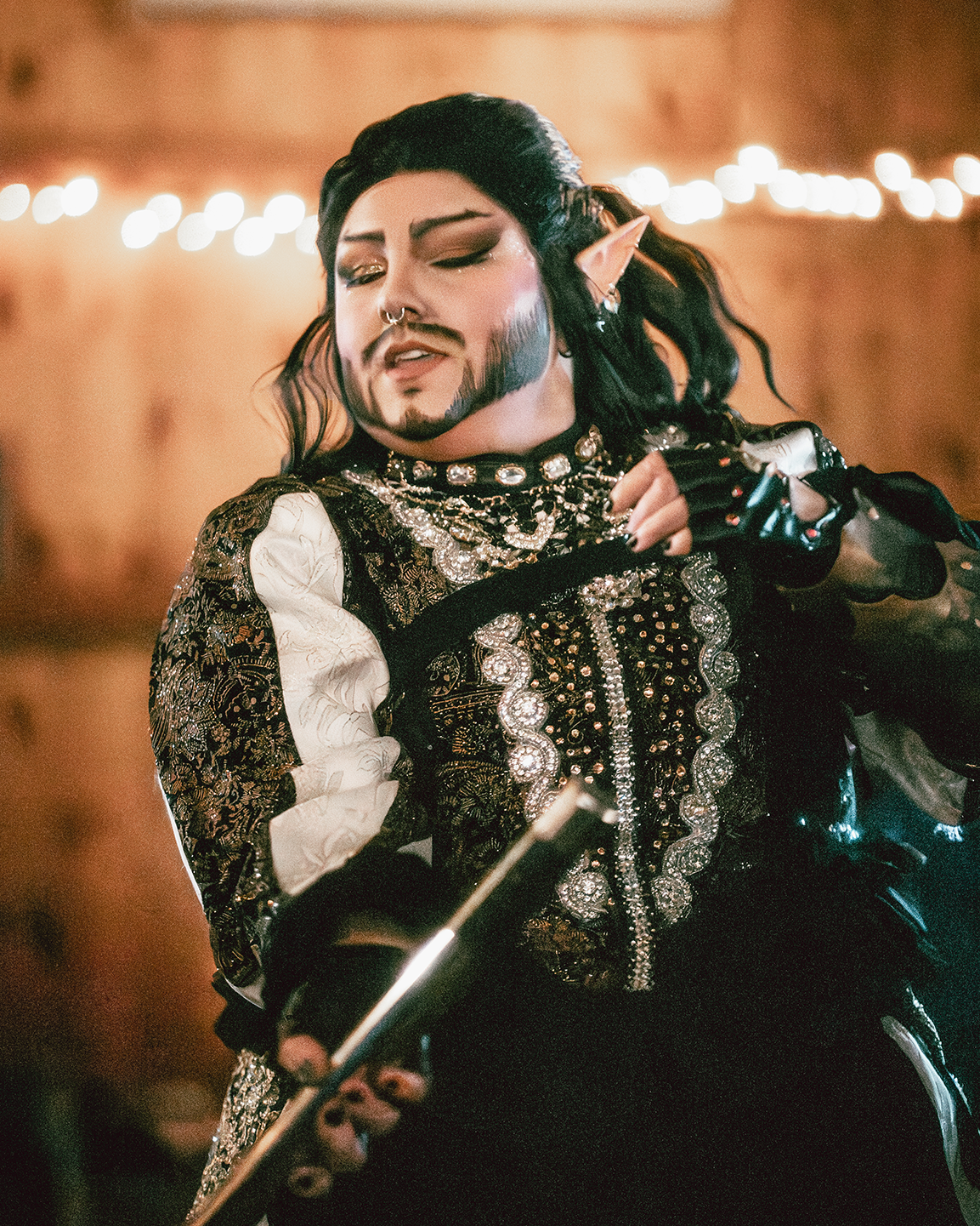 Person dressed in elaborate, dark, and ornate costume with jewelry, makeup, and elf-like ears, sitting on a motorcycle, with string lights in the background.