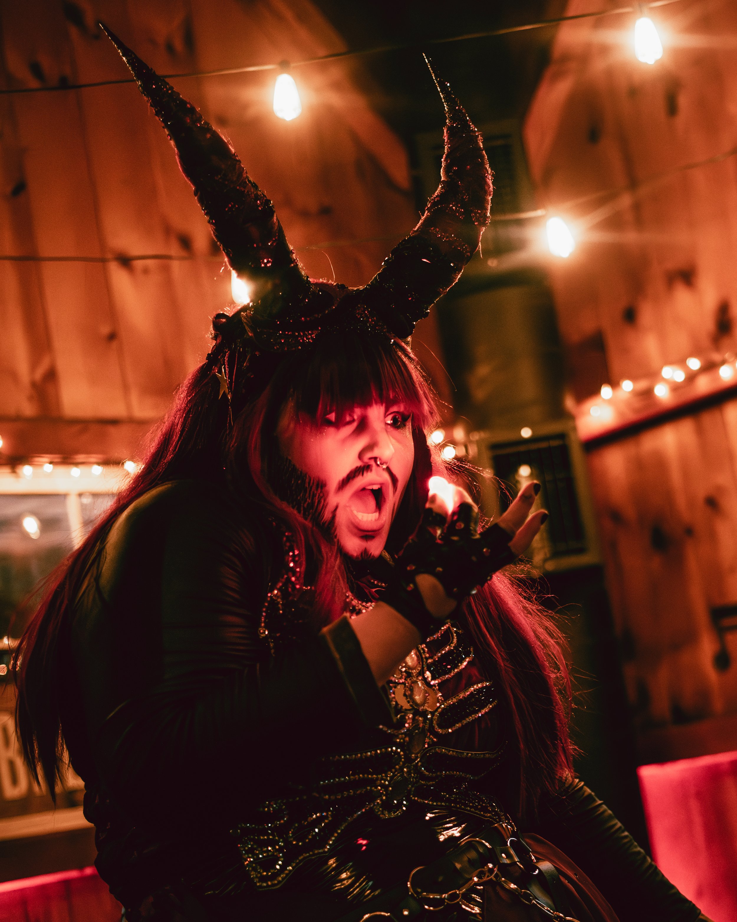 Person dressed in devil costume with black and red horns, in a room with warm string lights and wooden walls, making a dramatic gesture with a surprised or intense facial expression.