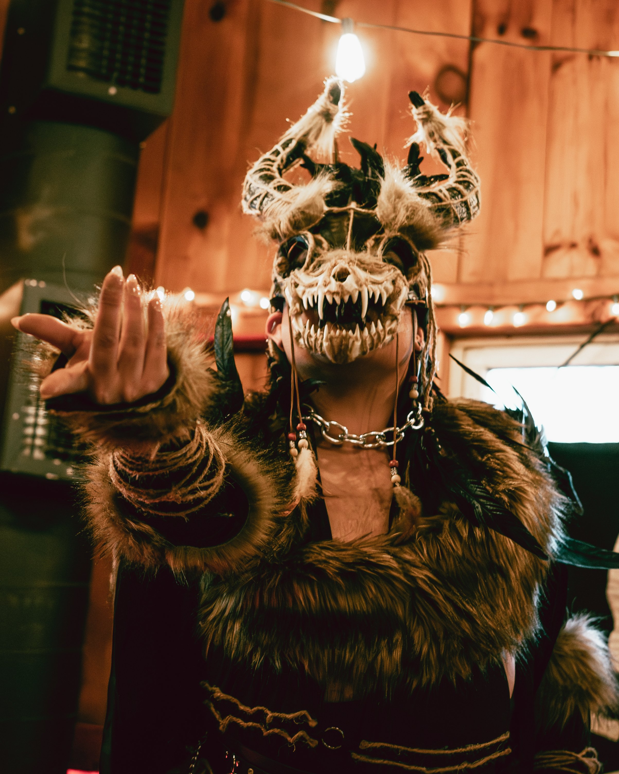 Person wearing a detailed animal skull and horns mask, with tribal jewelry and fur clothing, in a wooden room decorated with string lights.