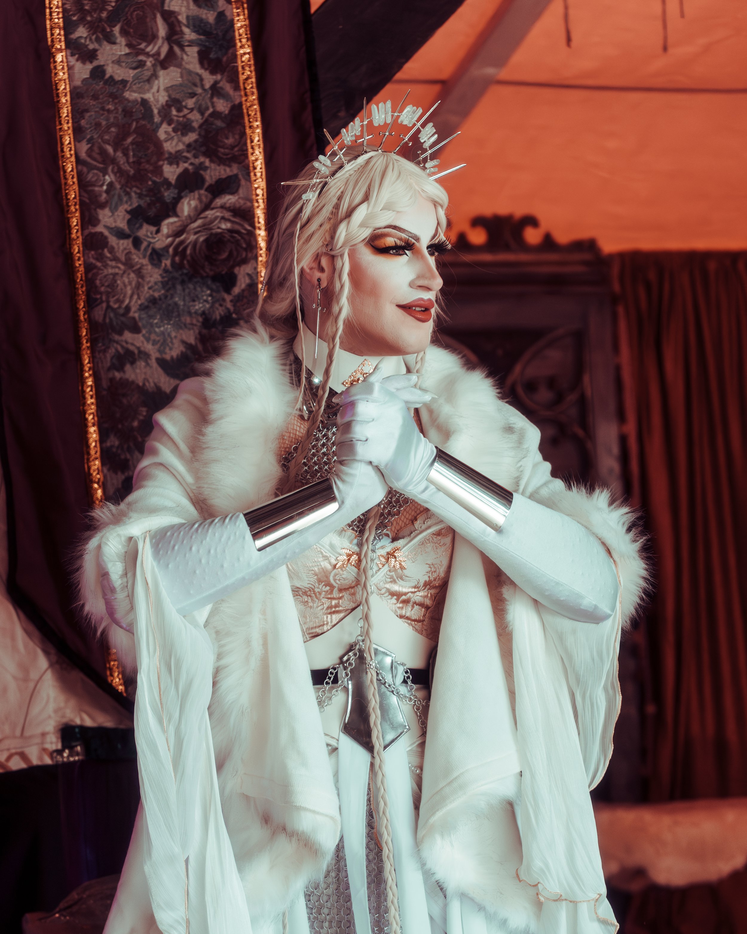 Person dressed in elaborate fantasy costume with long white gloves, fur cape, metallic accessories, and a silver crown with spikes, posing with hands clasped near face in a room with dark ornate curtains and warm lighting.