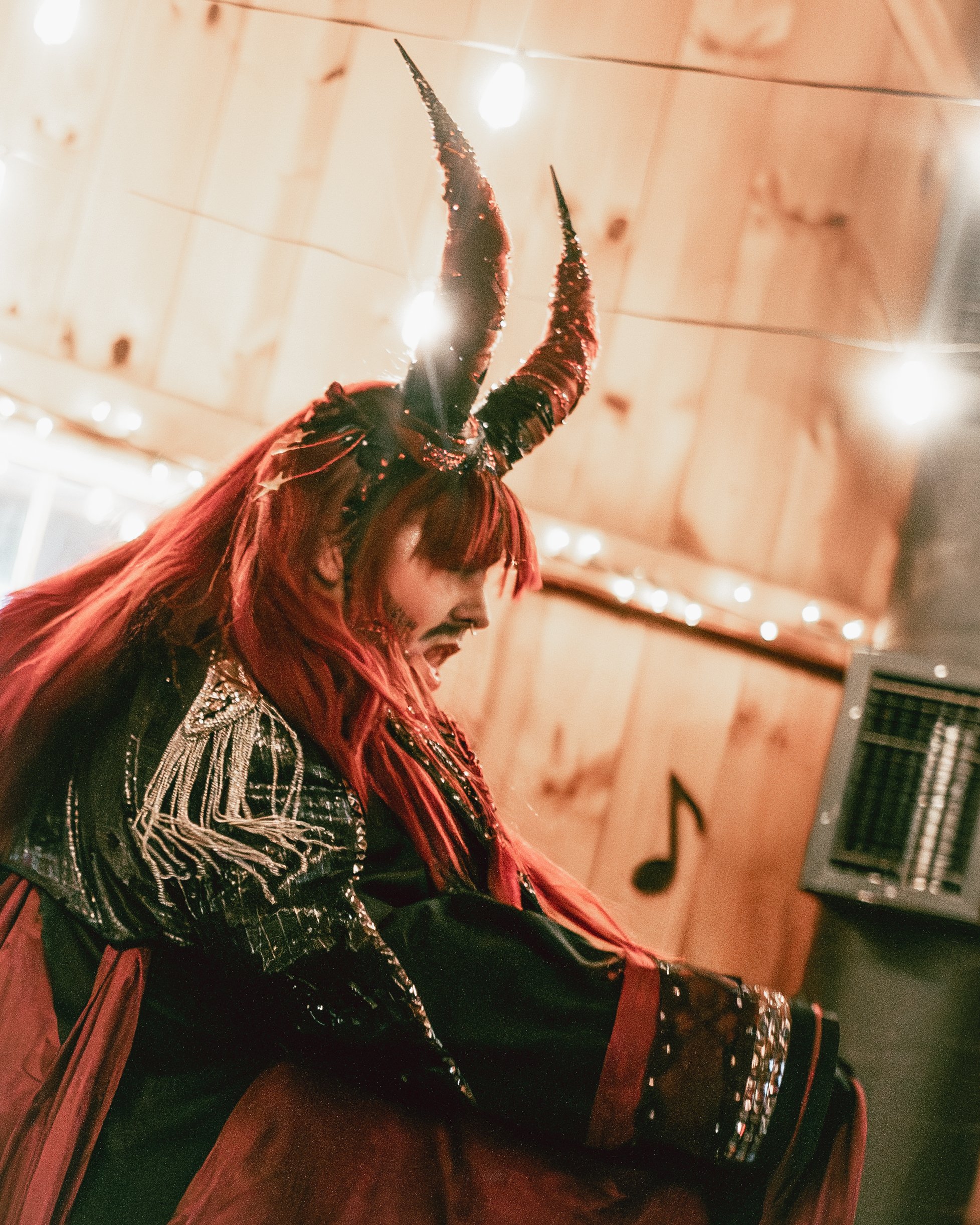 Person dressed in a costume with red hair, black and red clothing, and tall black and red horns, sitting and laughing or smiling, against a wooden wall with string lights.