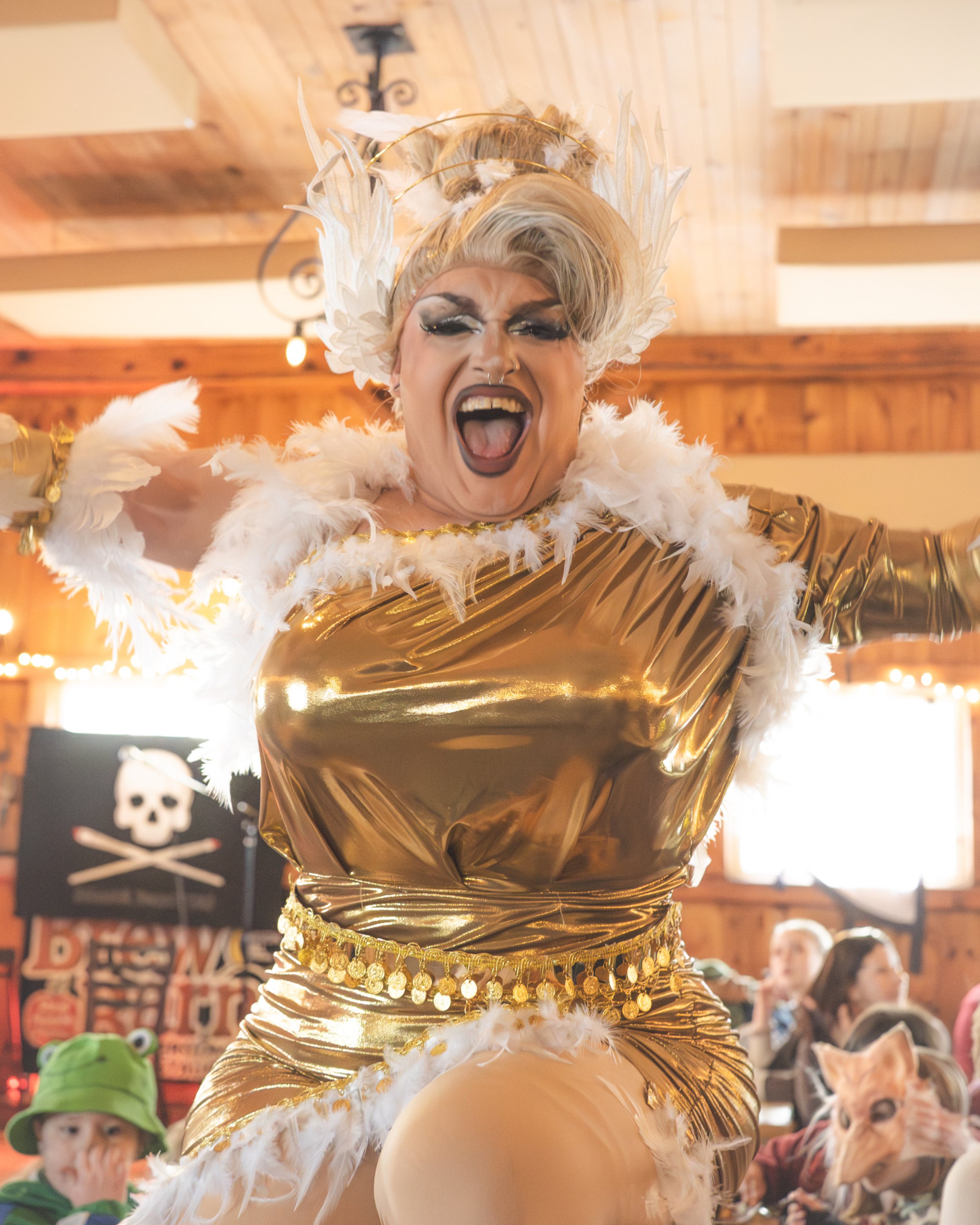 A person dressed in a shiny gold and white feathered costume with dramatic makeup, passionately singing or shouting at an indoor event.