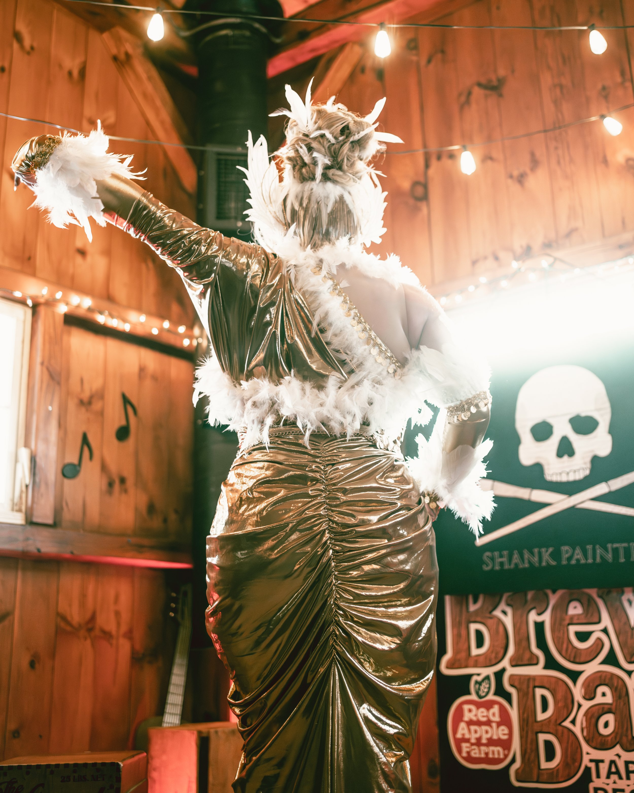 A person dressed in a shiny, gold gown with white feathers, standing inside a wooden-themed venue with string lights and a skull painting on the wall.