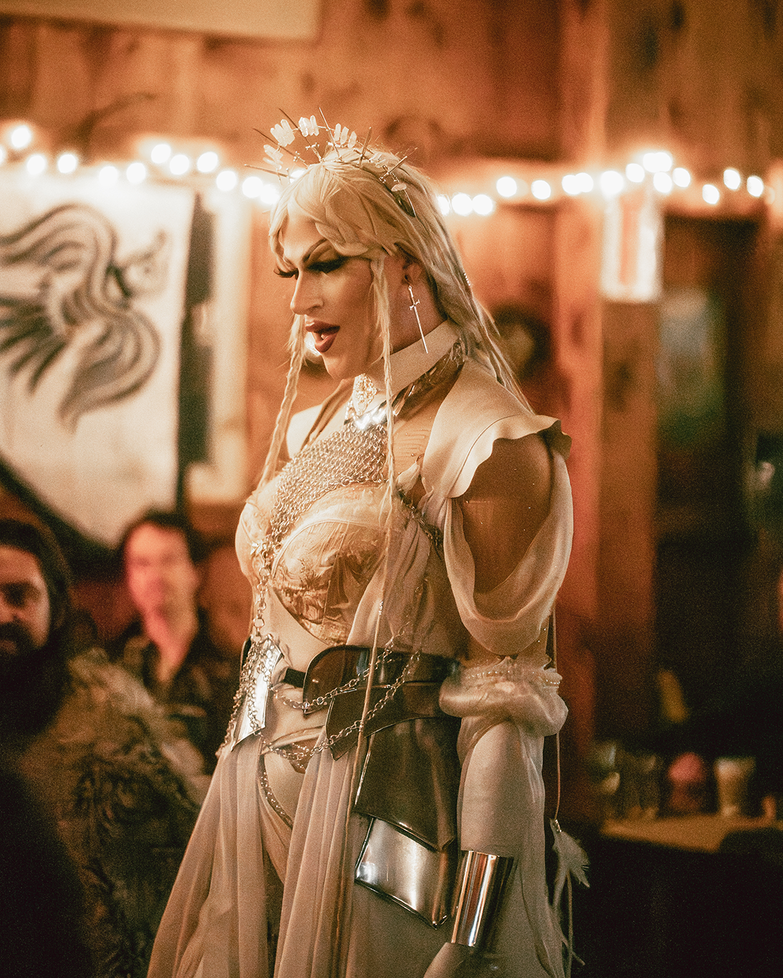 A person with platinum blonde hair styled with braids and a halo headpiece, wearing a beige and metallic outfit with chains, standing in a dimly lit, rustic indoor space decorated with string lights, with people sitting in the background.