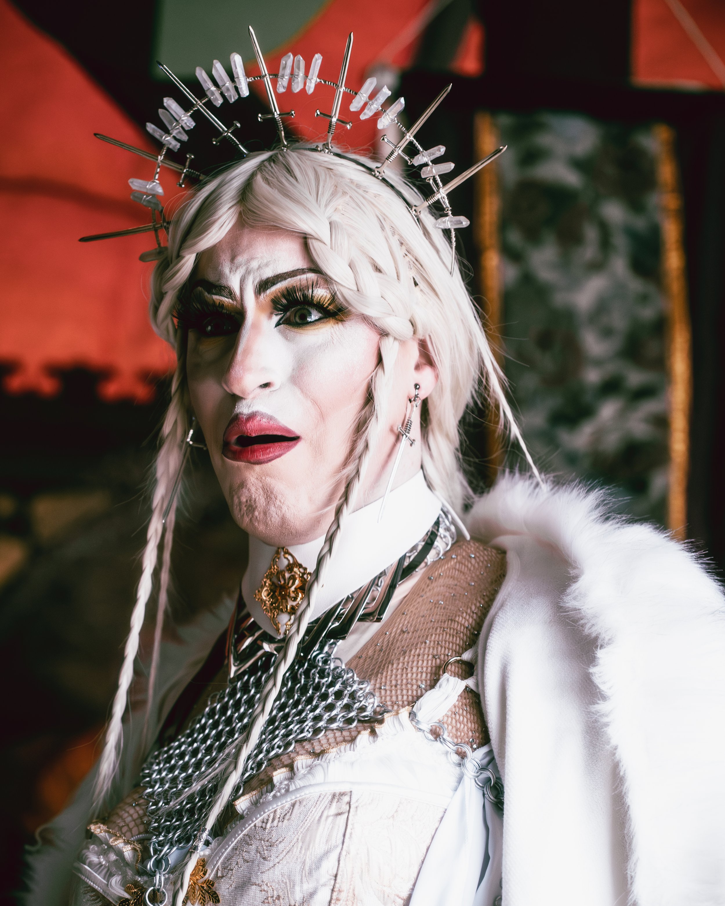 Person dressed as a queen with platinum blonde hair, wearing a crown made of metal and crystals, in a regal costume with jewelry and a white cape.
