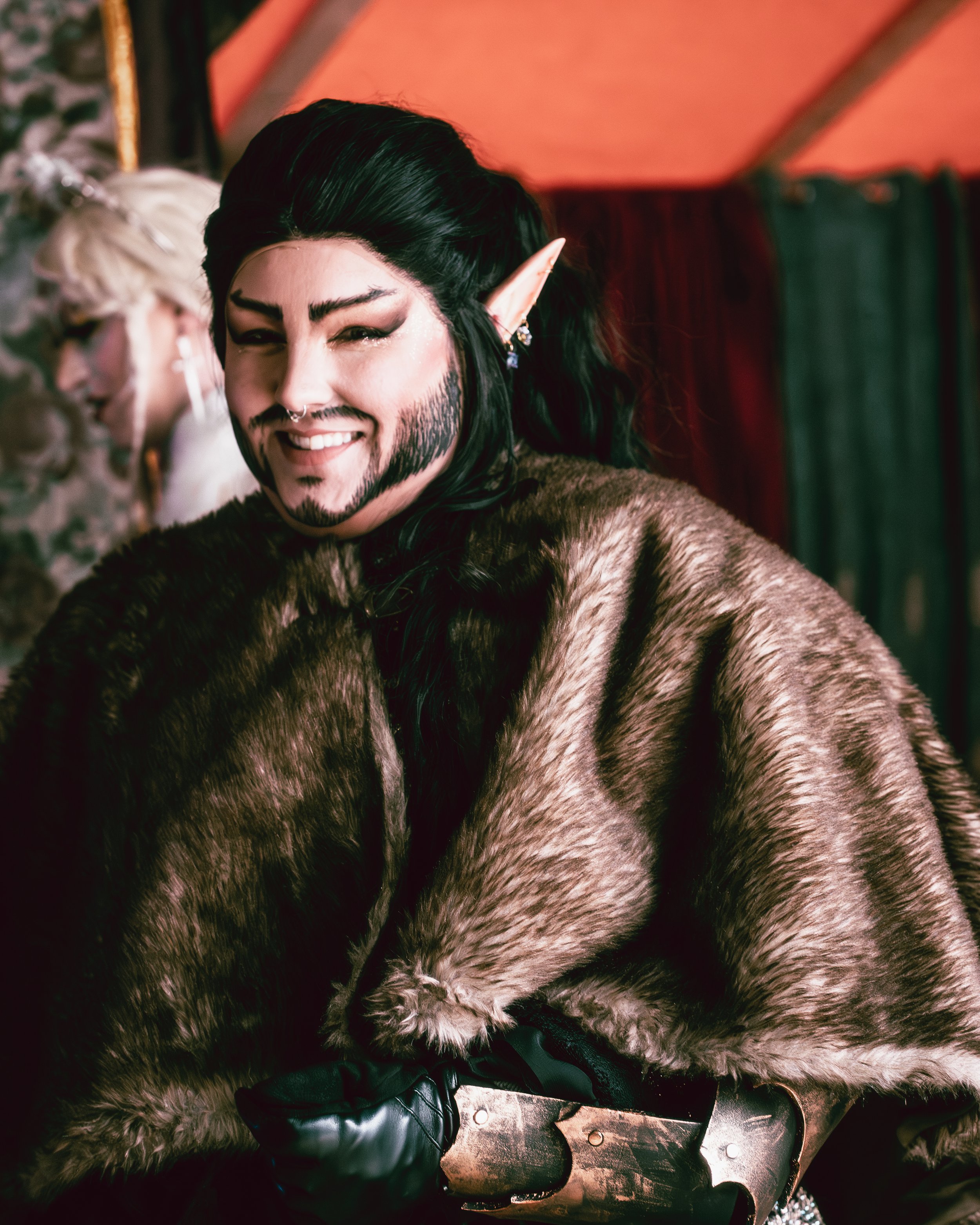 Person dressed as an elf with long black hair, a beard, a faux fur cape, and a leather glove, smiling at the camera.