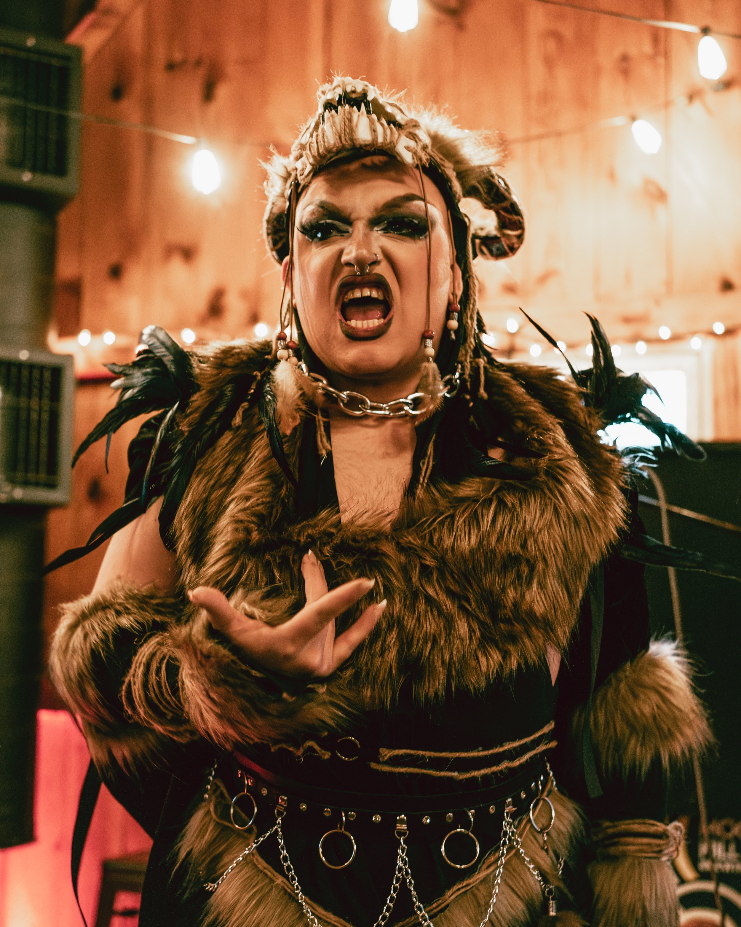 A person dressed in a fierce, tribal-inspired costume with fur and feathers, wearing a headpiece resembling an animal skull, making an aggressive facial expression with mouth open, showing teeth, and gesturing with one hand. The background features w