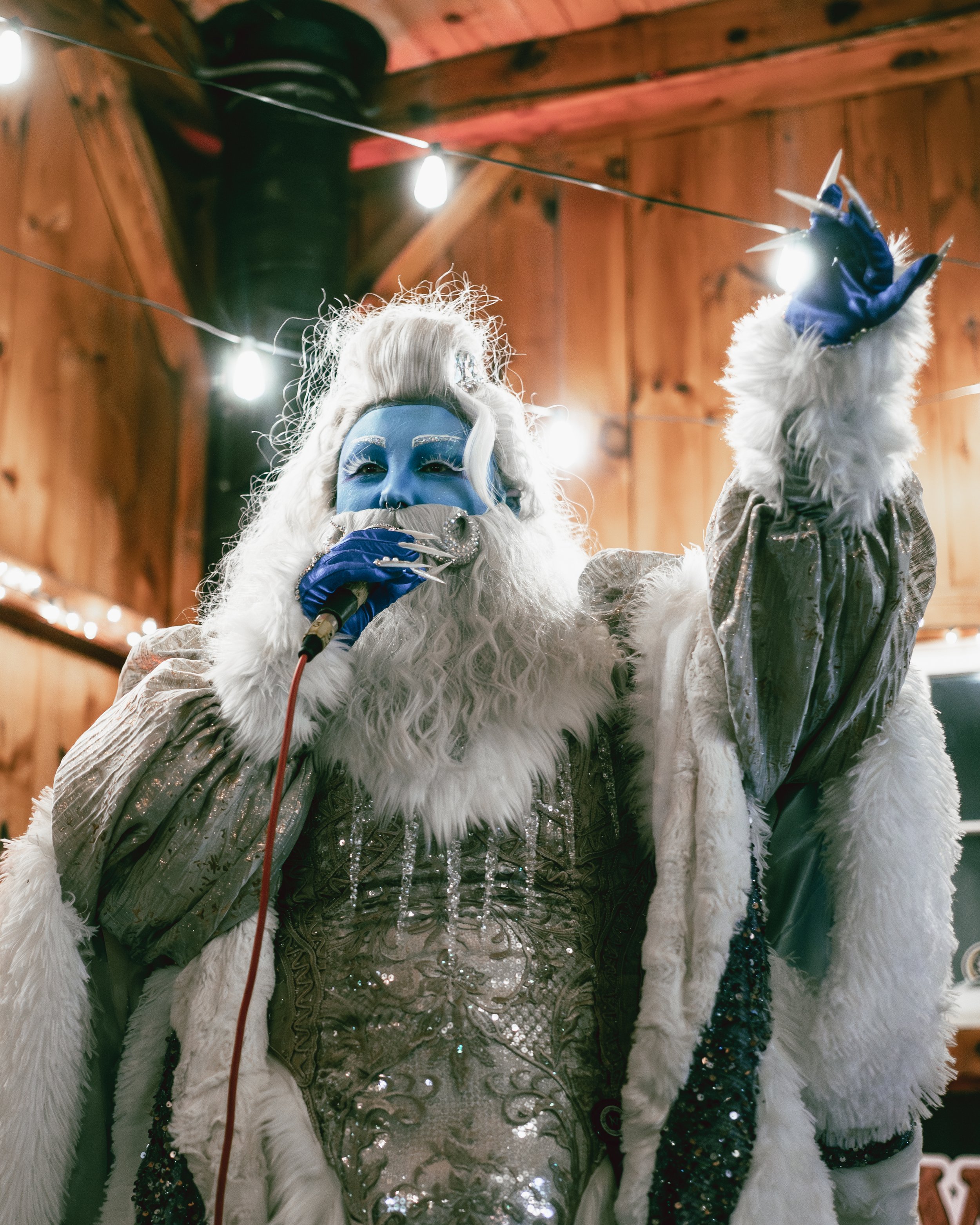 Person dressed as the Ice Queen from 'Frozen,' with blue face paint and elaborate costume, singing into a microphone in a wooden, festively decorated setting.