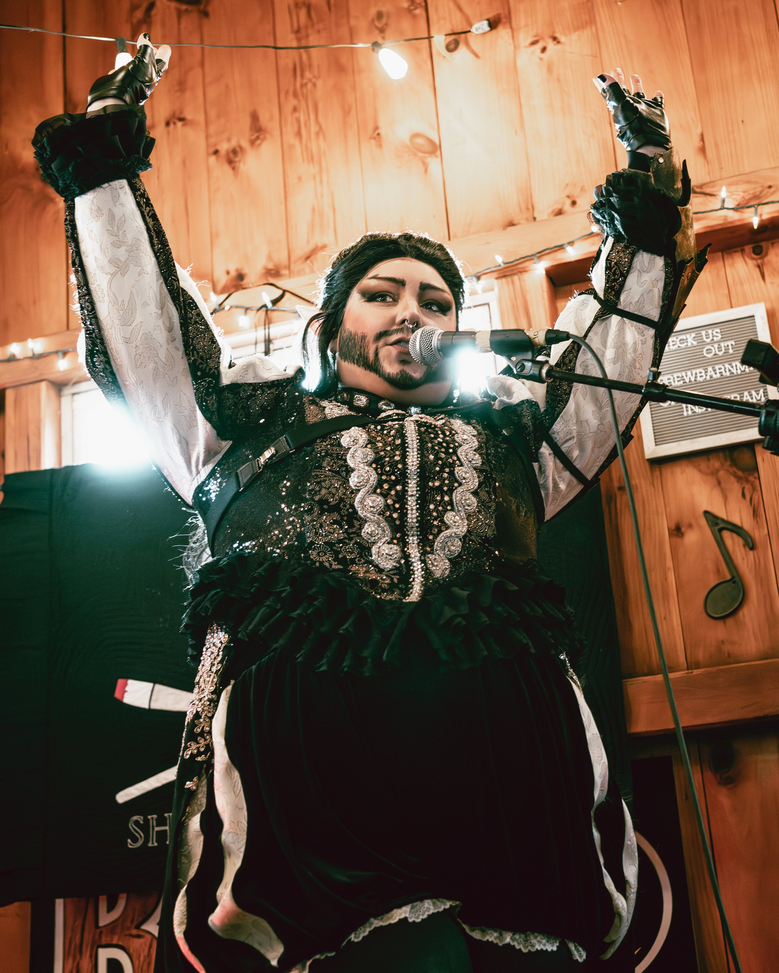 A woman dressed in gothic and vintage-style clothing stands on stage, singing into a microphone with her arms raised. She has dark hair, makeup, and face paint, with a wooden wall and string lights in the background.