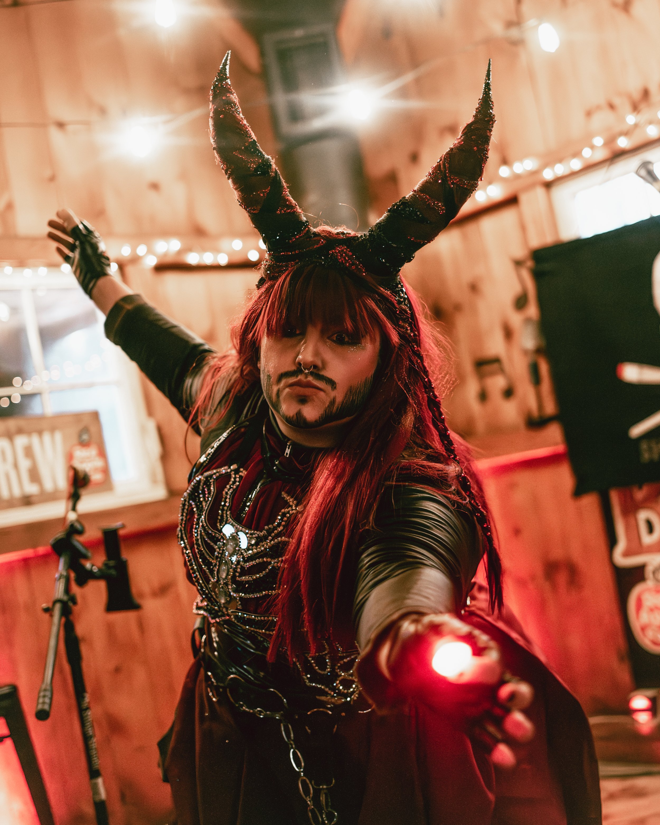 Person dressed as a demon with black and red horns, dark makeup, and long red hair, holding a small red light in a dimly lit wooden room decorated with string lights.