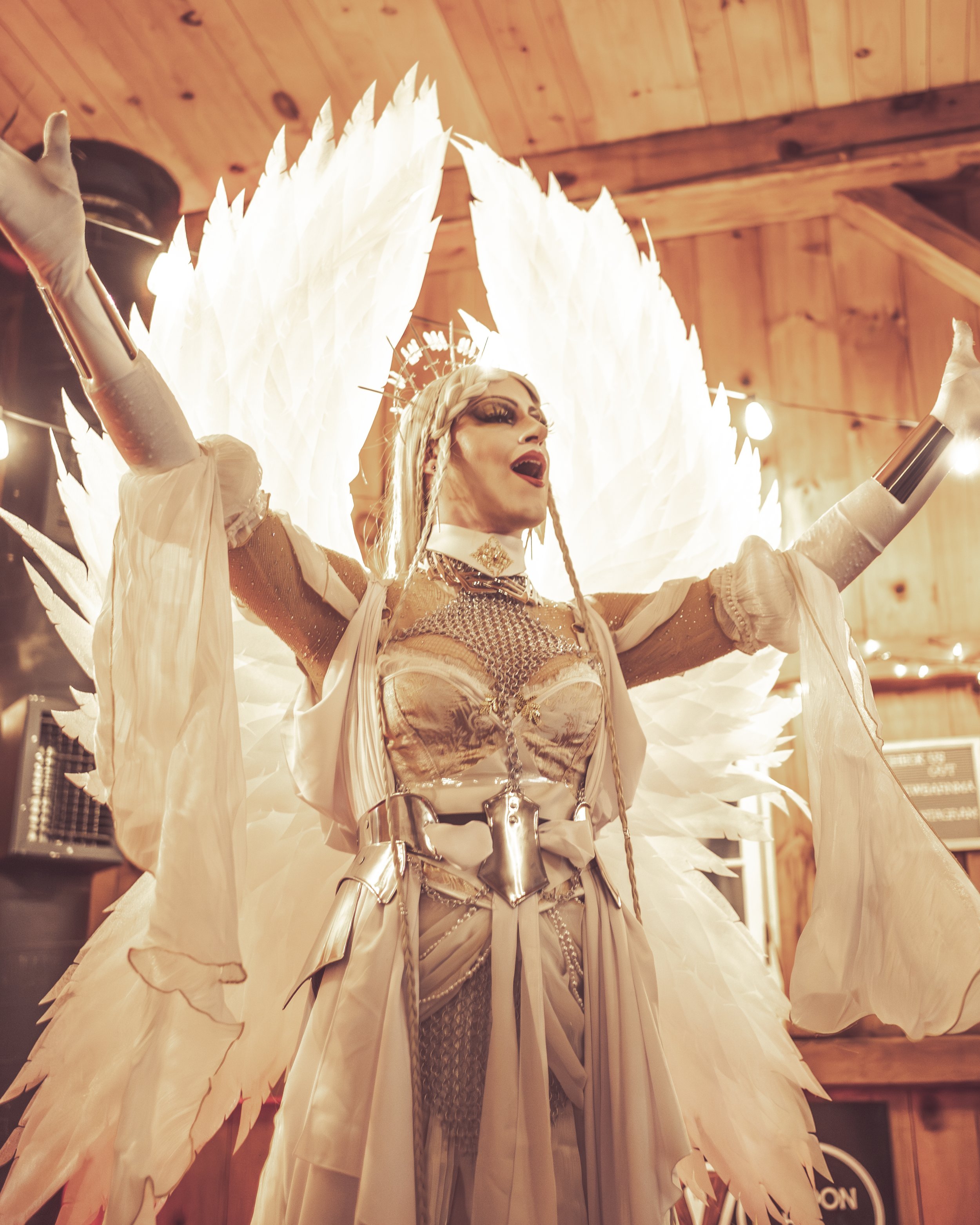 Performer in elaborate costume with large white feathered wings and crown, singing with arms raised in a wooden venue.