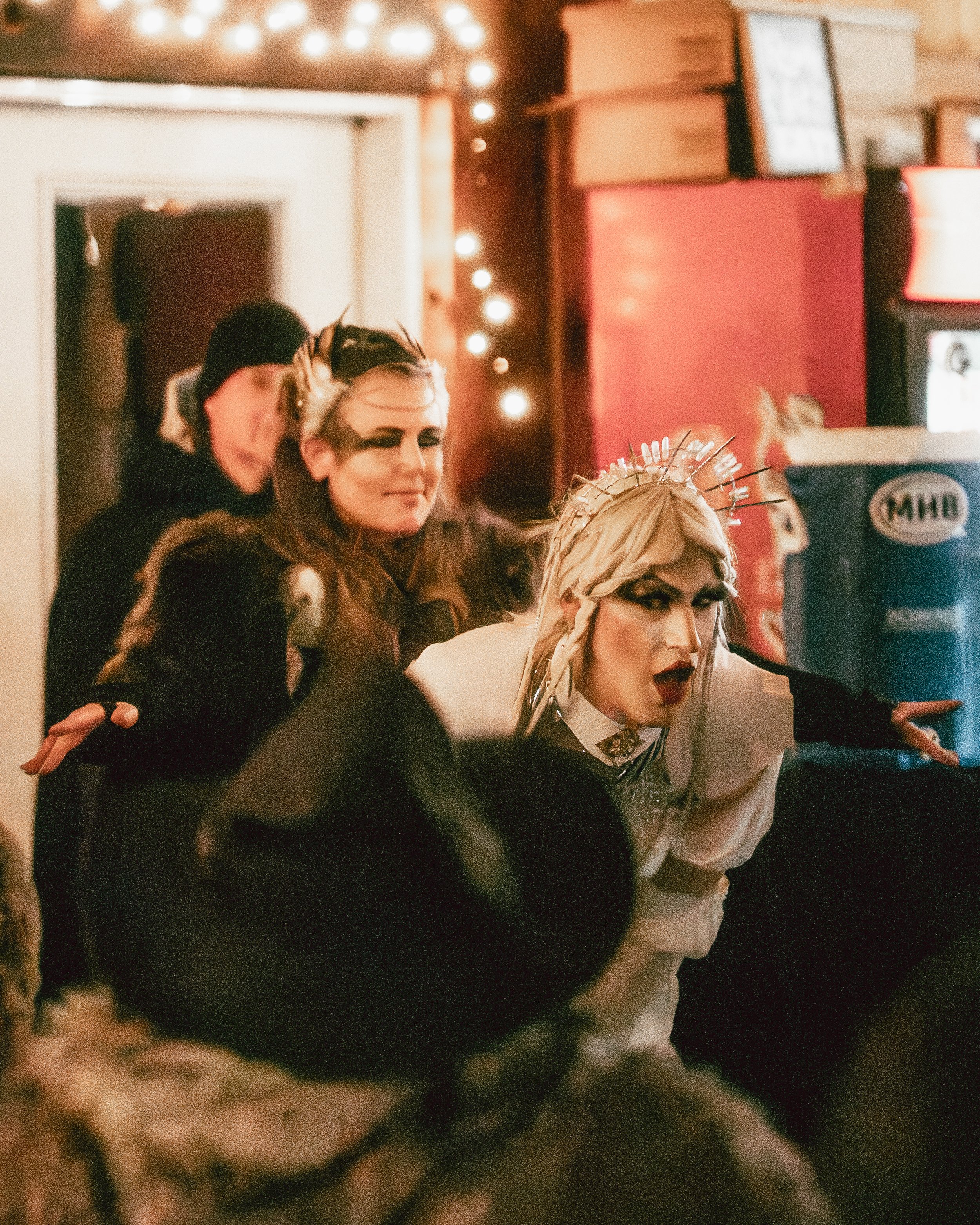 Three women with elaborate makeup and costumes perform at a lively indoor event. One woman has platinum blonde hair and a crown-like headpiece.