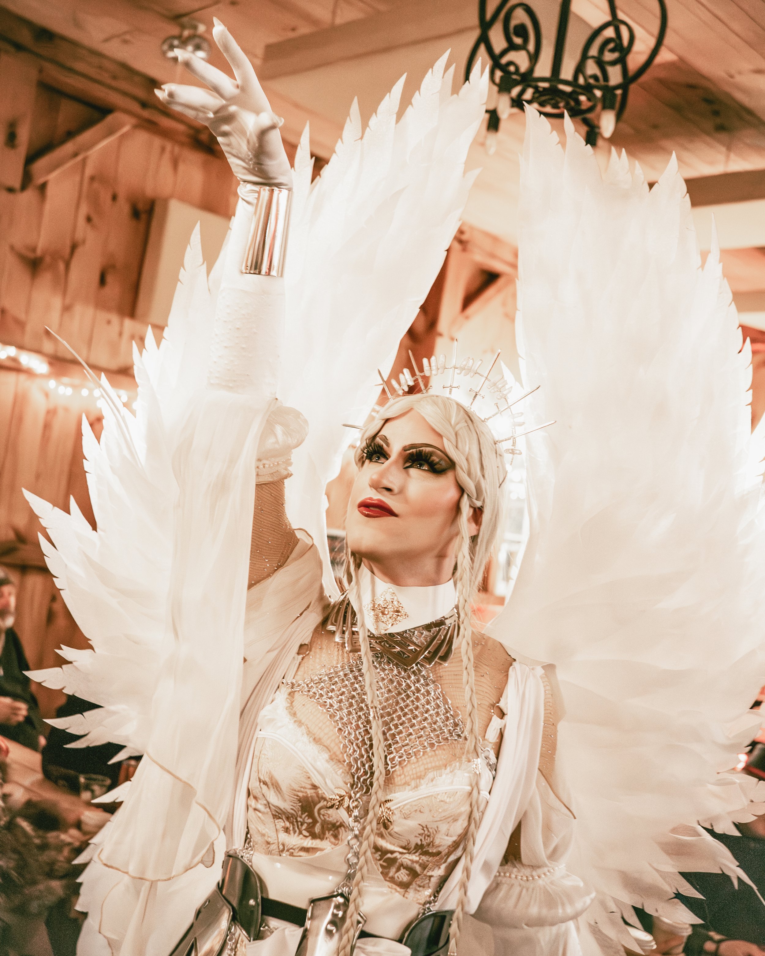 A person dressed in an elaborate angel costume with large white feathered wings, wearing a crown, makeup, and light-colored clothing, raising one arm in a celebratory gesture.