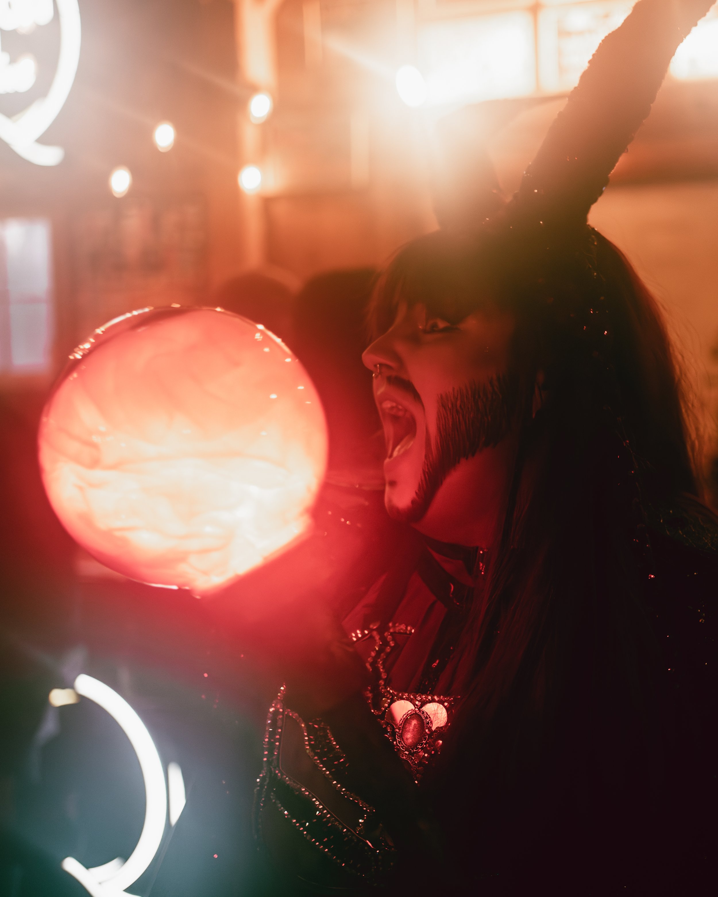 A person with clown makeup and black face paint, wearing a costume with glitter, is about to bite or lick a glowing orange orb, with their mouth open wide.