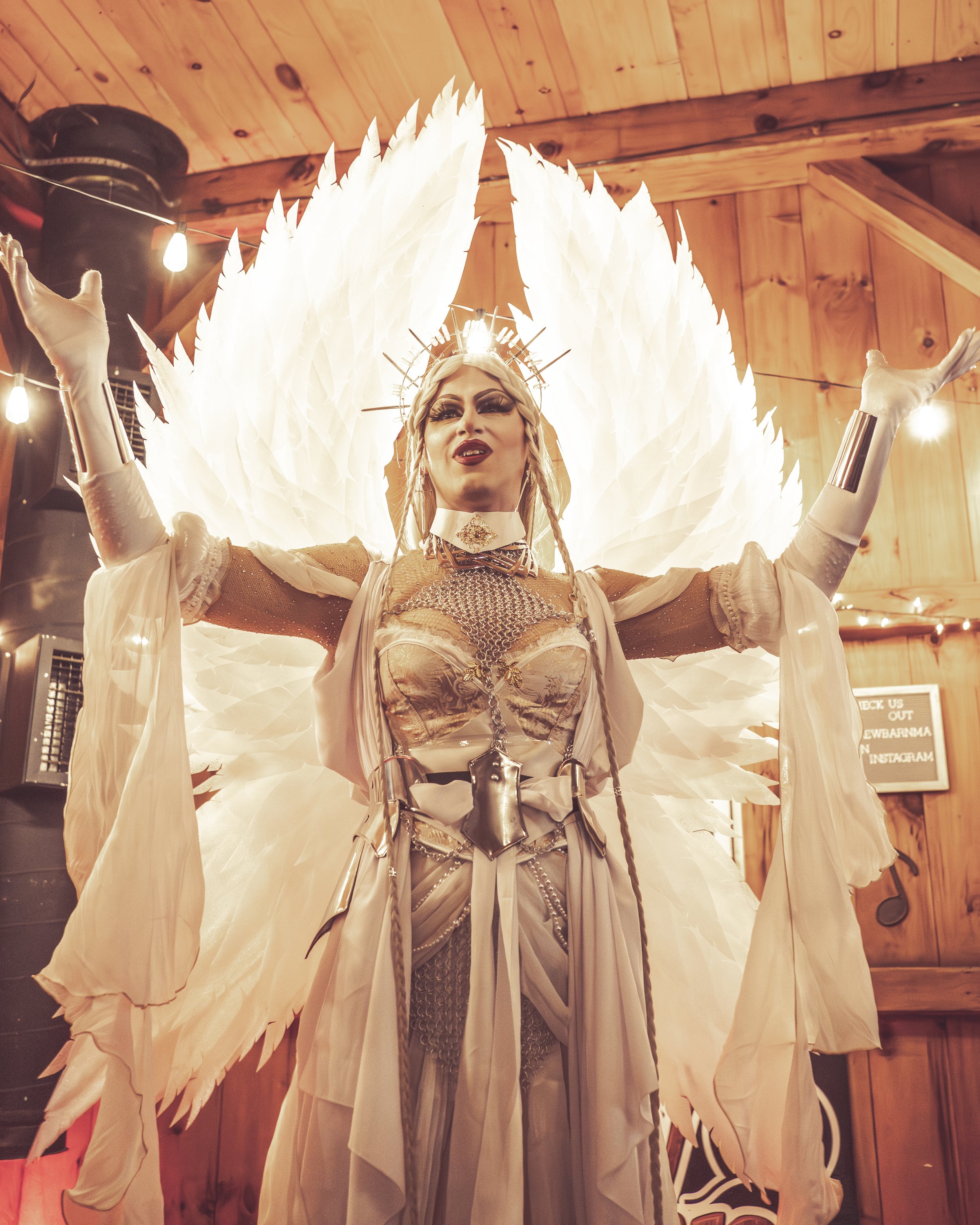 A drag performer dressed as an angel with large illuminated wings, wearing elaborate makeup and a metallic and white costume, in a wooden indoor setting with string lights.