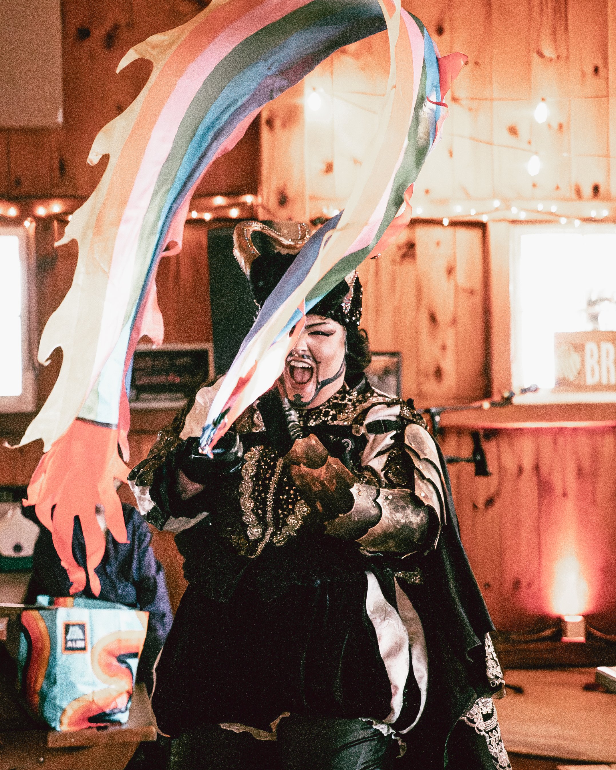 Woman dressed in an elaborate costume, holding colorful streamers and a sword, expressing excitement or joy in a warmly lit, wooden-paneled room decorated for a celebration.