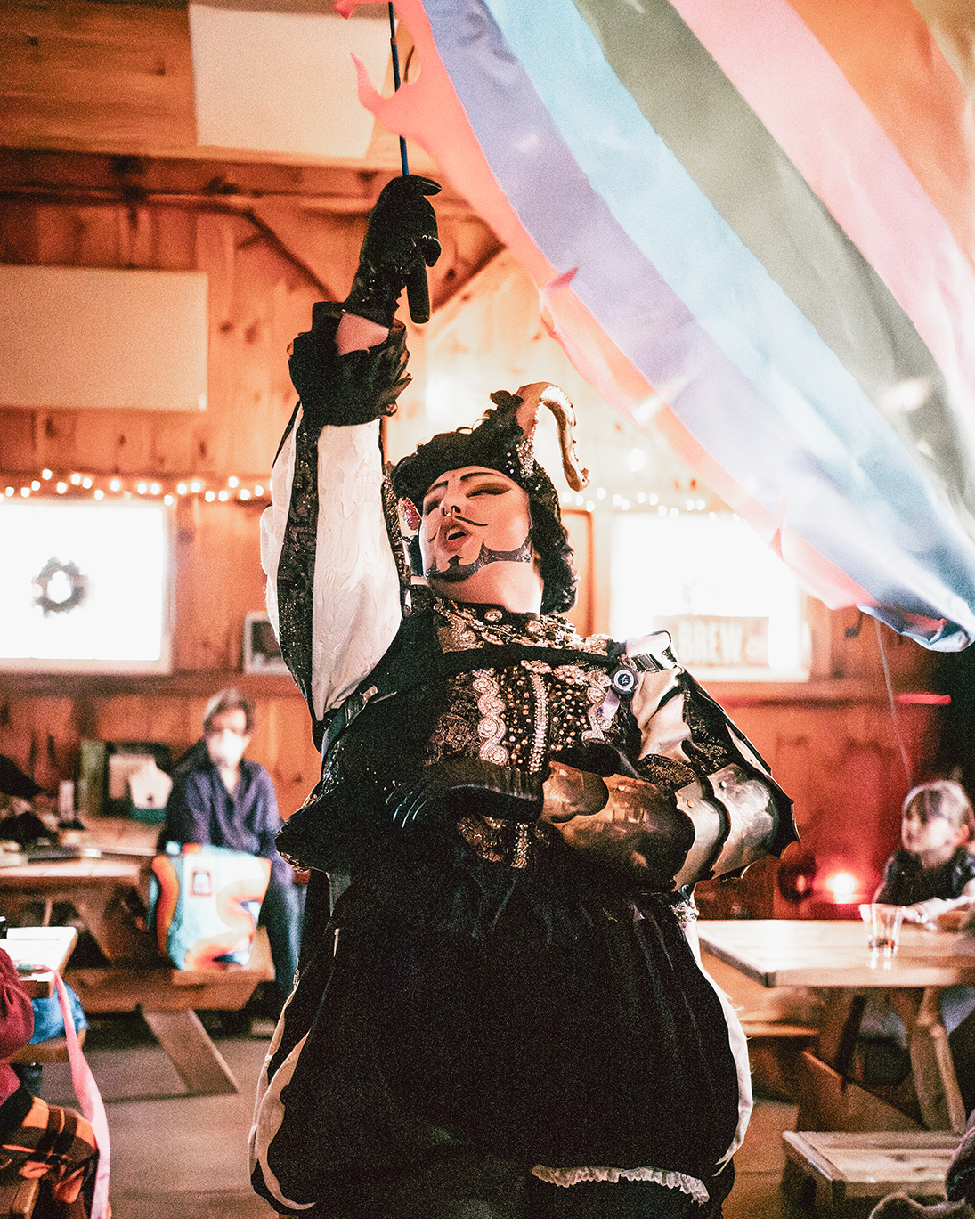 A person dressed in an elaborate costume with horns and facial makeup holding colorful streamers or decorations in a festive indoor setting.