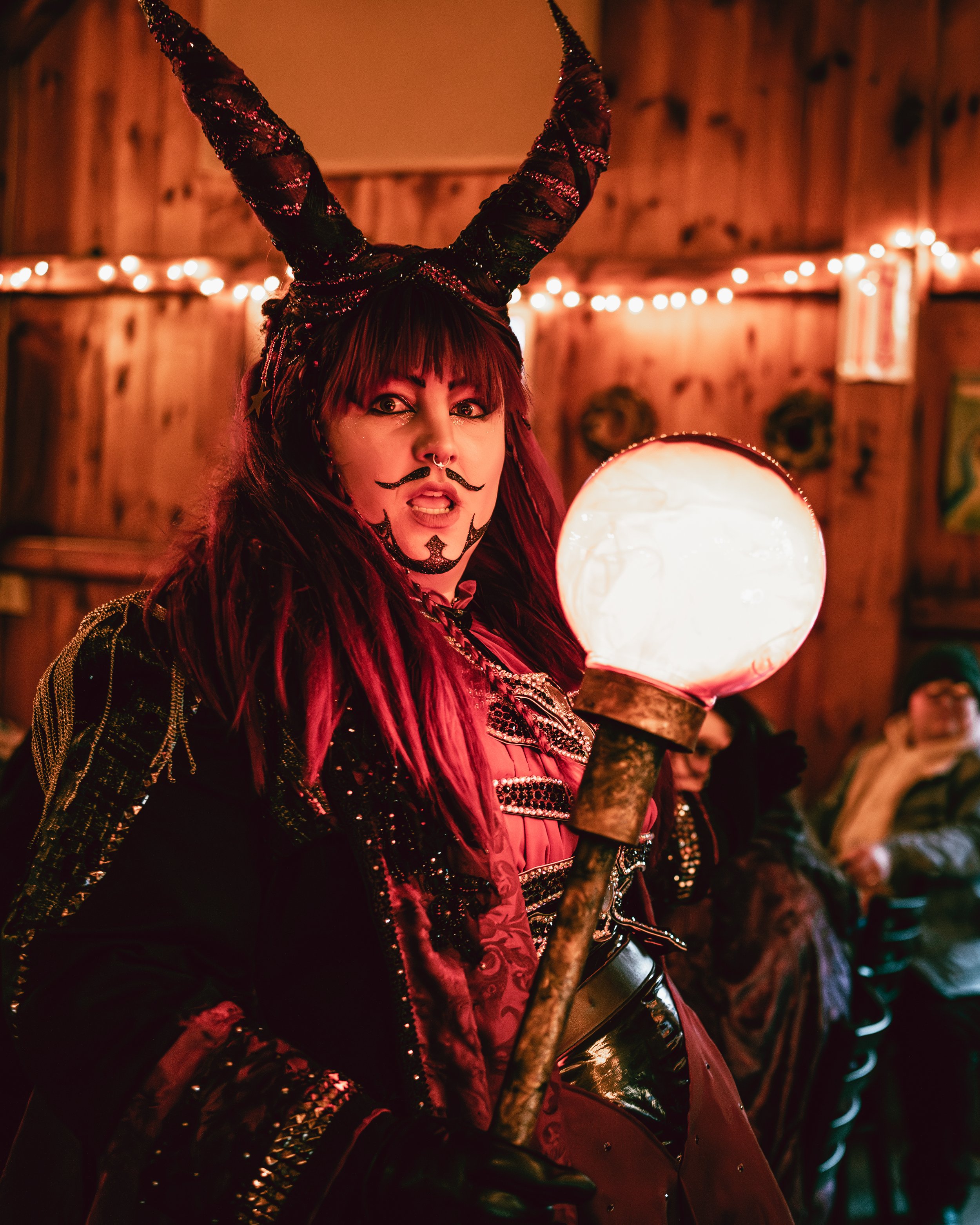 Person in a fantasy costume with devil-like horns, face paint, and a staff with a glowing orb, in a decorated indoor setting.