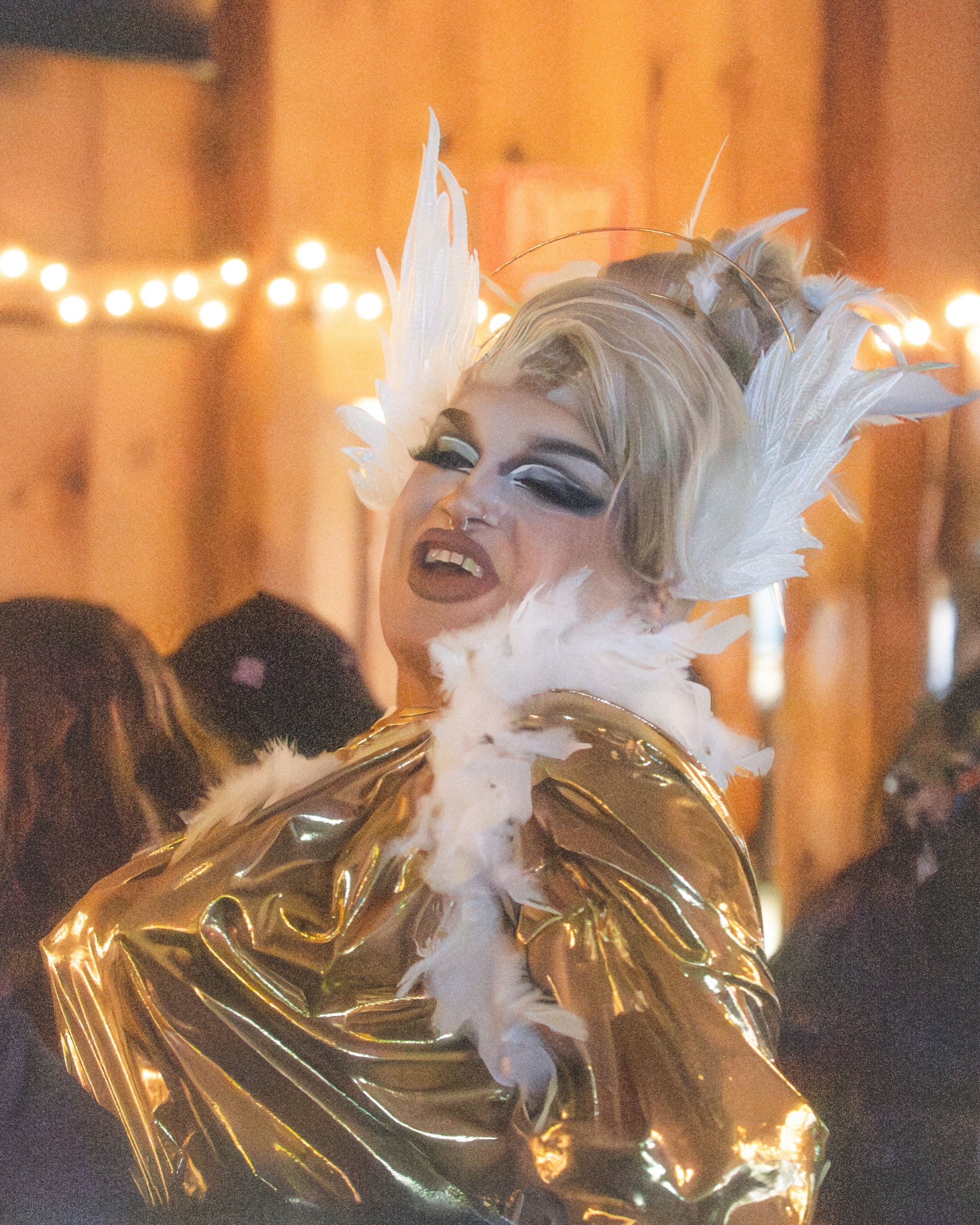 Person dressed in a dramatic, fashionable outfit with feathered headpiece and metallic gold clothing, performing with an expressive facial expression in a warmly lit indoor setting.