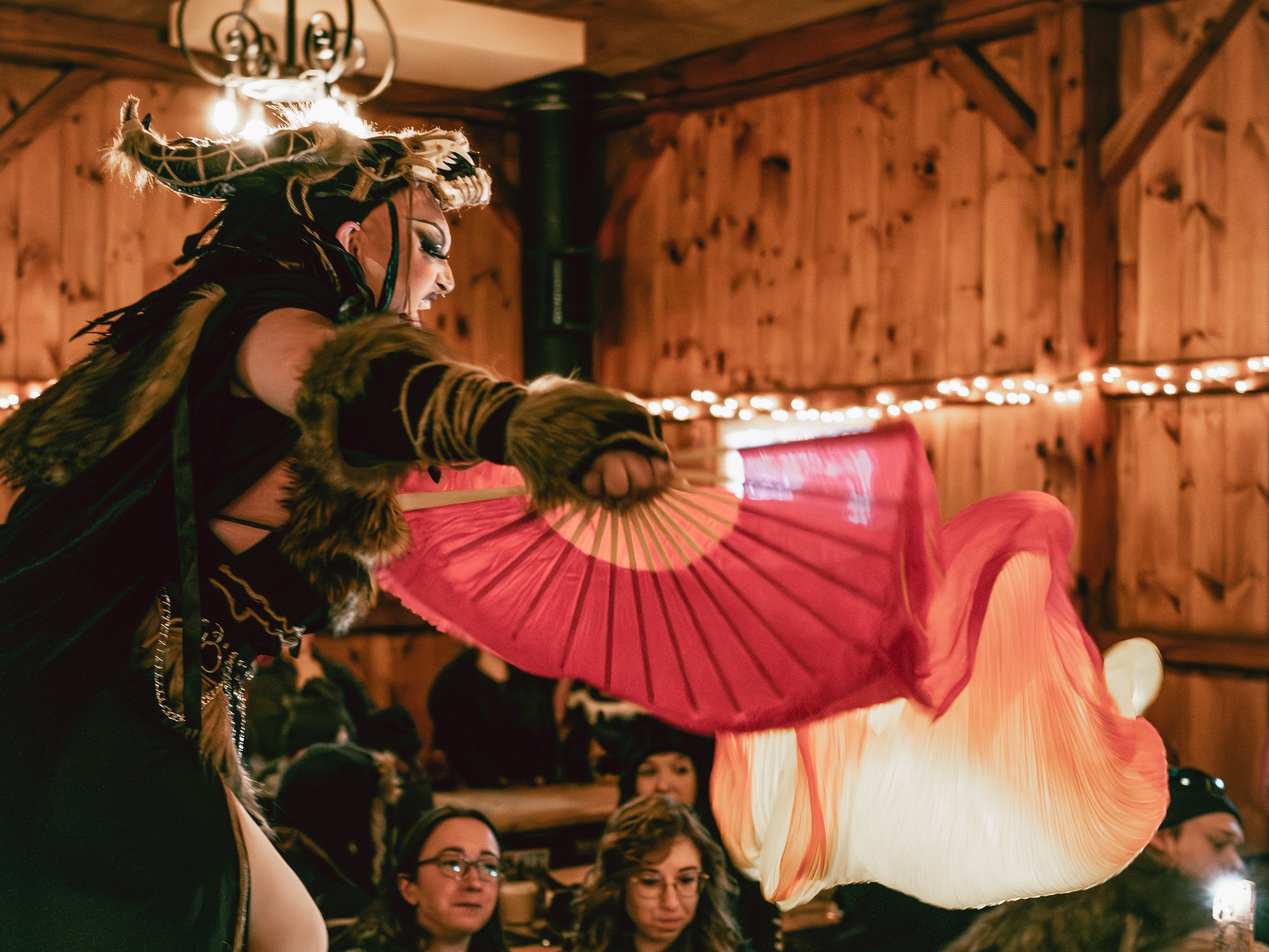 A performer dressed in a costume with fur and a headpiece with horns is spinning a large pink and white silk fan during a performance in a wood-paneled room with string lights.