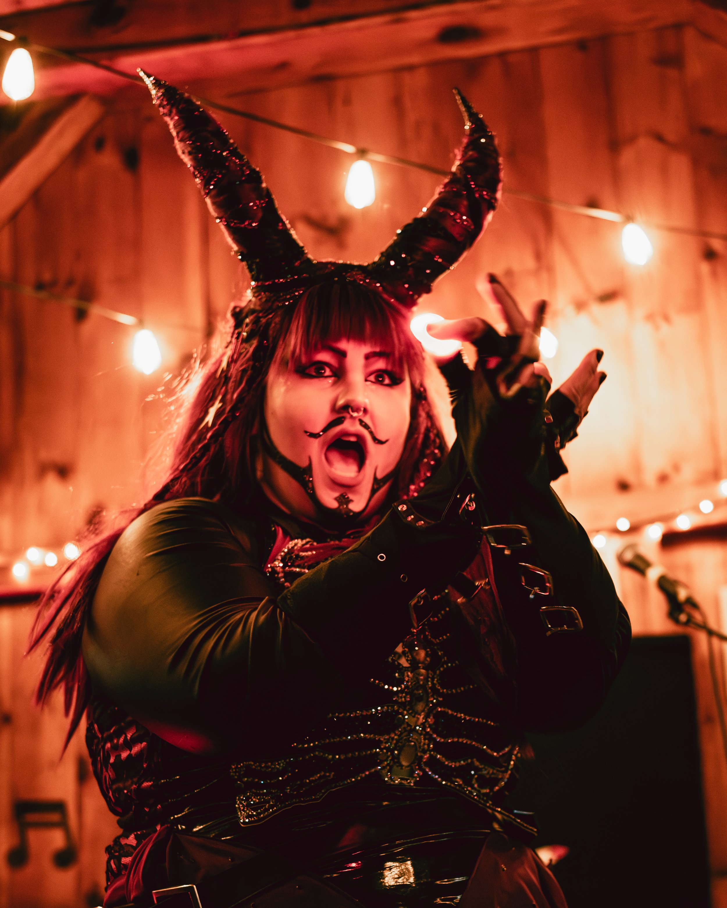 A person in a devil costume with black and red horns, makeup, and face paint, singing or performing on stage with wooden walls, string lights, and microphones in the background.