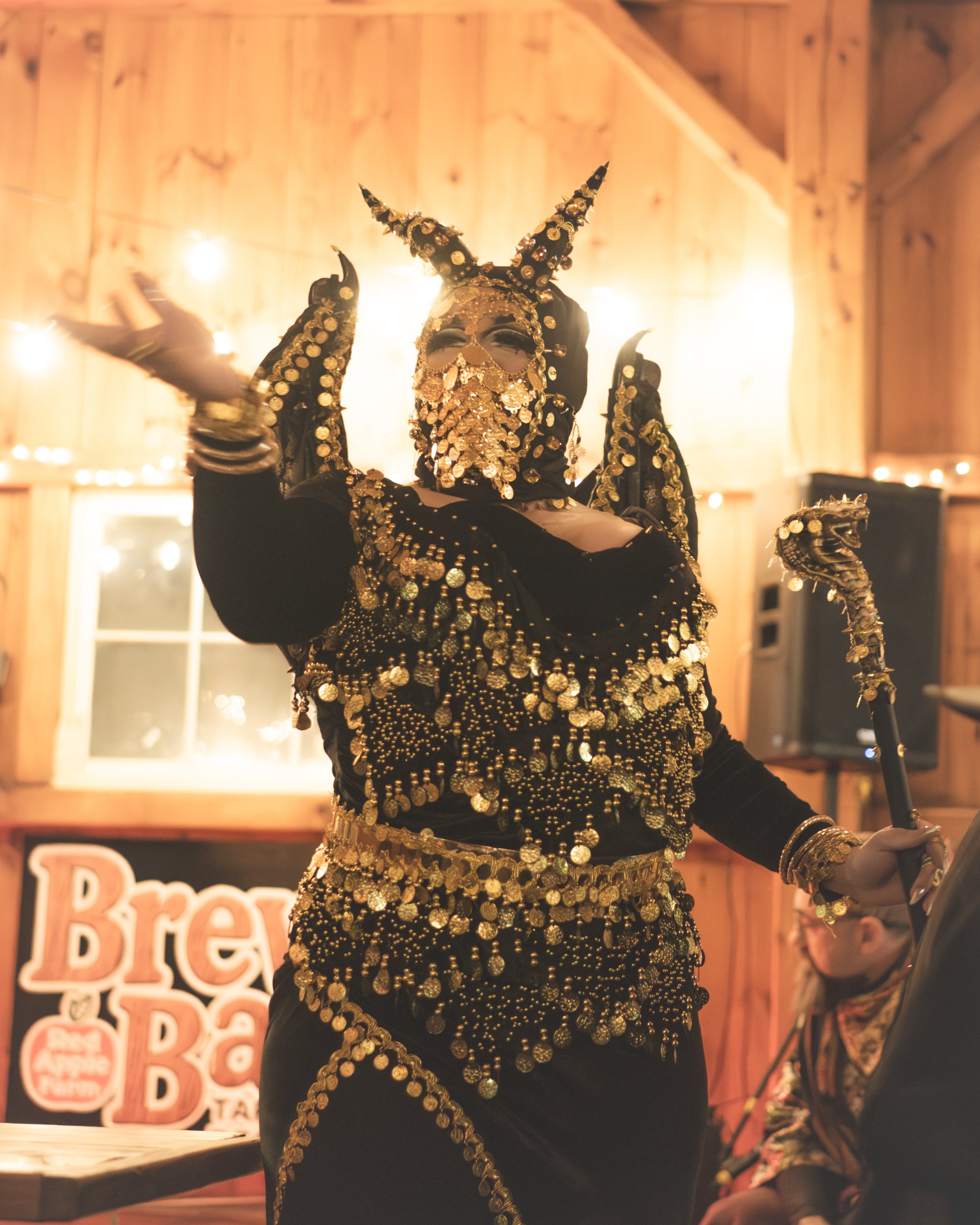 Person wearing an elaborate black and gold costume with a horned mask, adorned with gold coins and jewelry, in a wooden room with string lights.