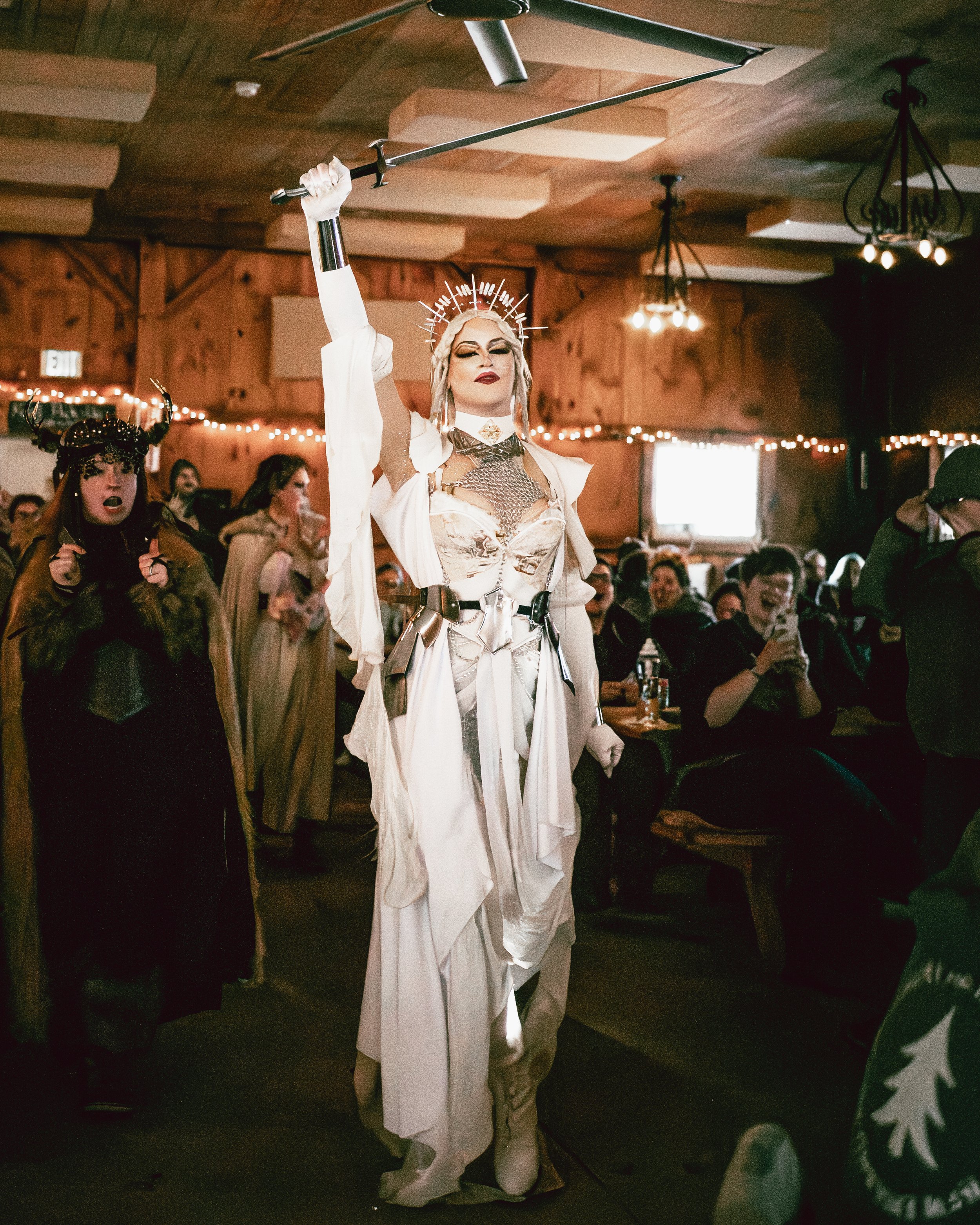 A person dressed in a dramatic, elaborate costume with a crown-like headpiece, holding a sword raised, standing in a room with wooden walls and string lights, surrounded by seated and standing attendees.