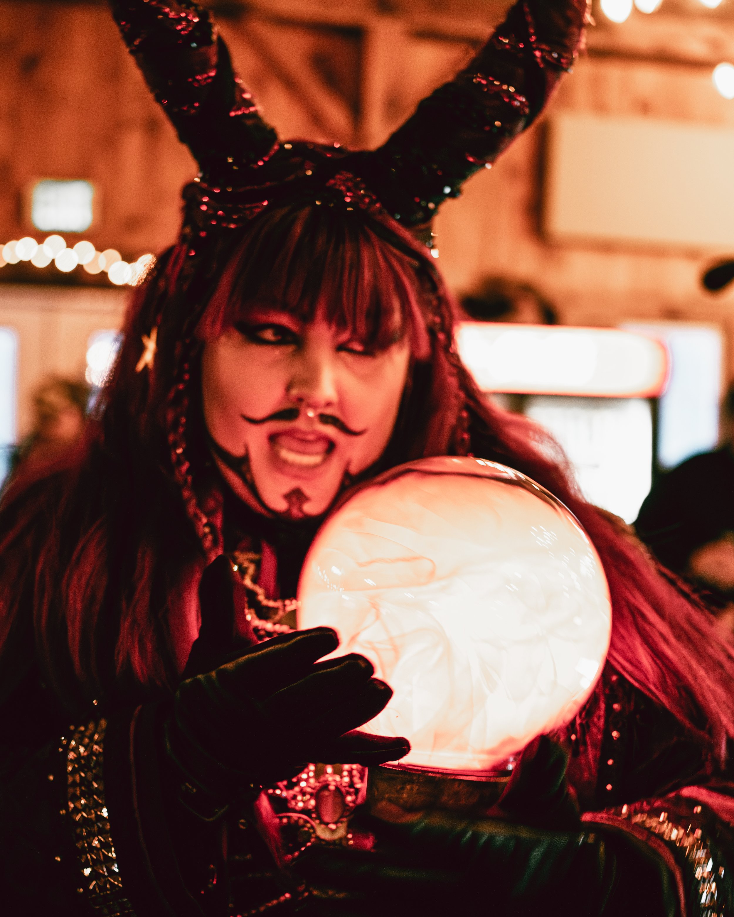 A person dressed in a black sequined costume with large horn-like headpieces holds a glowing crystal ball at a festive event, with blurred background lights and a lively atmosphere.