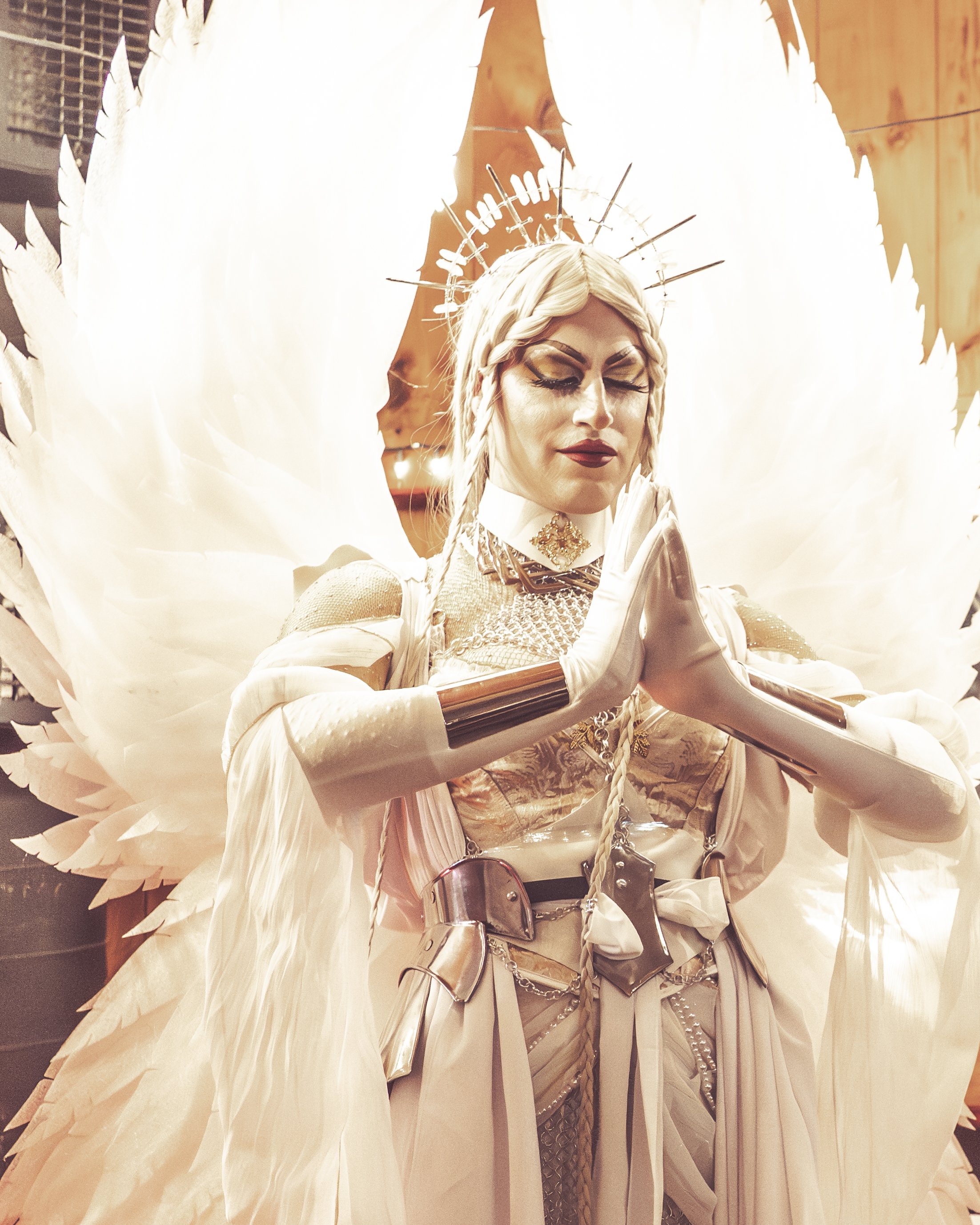 A woman dressed as an angel with white wings, wearing silver and white costume, with her hands pressed together in a prayer position and eyes closed.