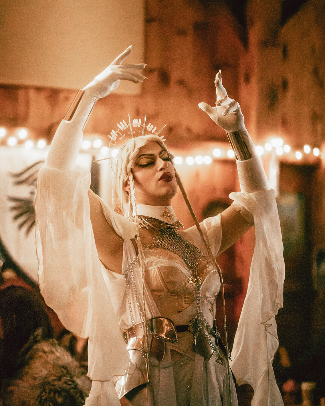 A person dressed in elaborate, futuristic costume with metallic accessories and dramatic makeup, posing with arms raised in a performance setting with warm lighting and string lights.