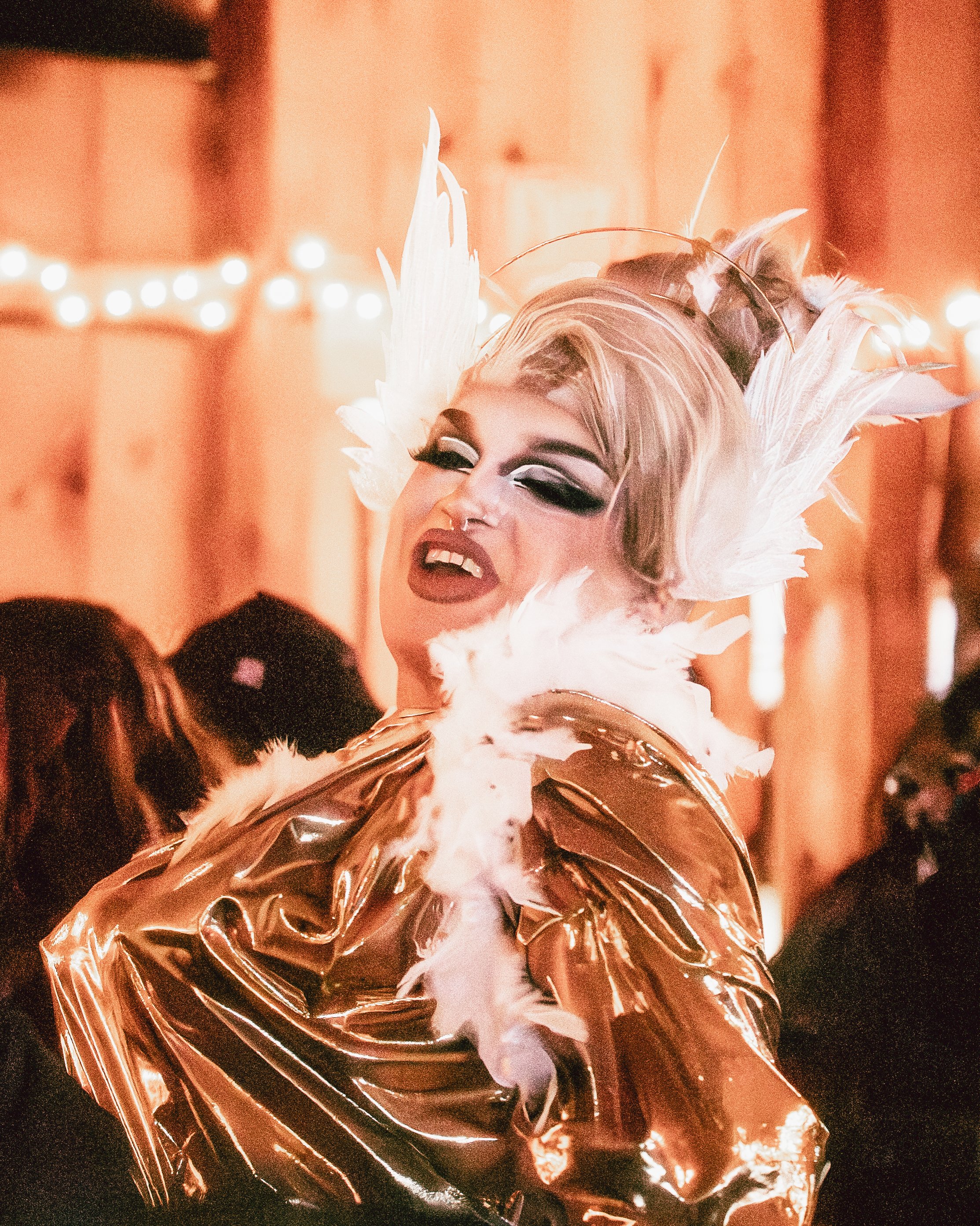 A drag queen with platinum blonde hair styled in an updo, wearing dramatic makeup with black eyeshadow, dark lipstick, and false eyelashes, dressed in a shiny metallic gold jacket with white feathers on the shoulders, in a lively party setting with b