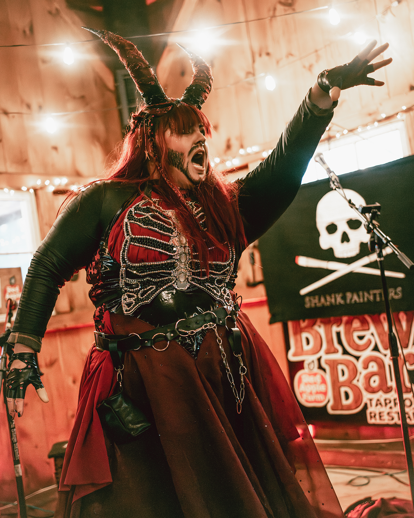 Person dressed in a dark, embellished costume with horns on their head, singing or shouting passionately on stage in a wooden venue with string lights and a skull banner in the background.