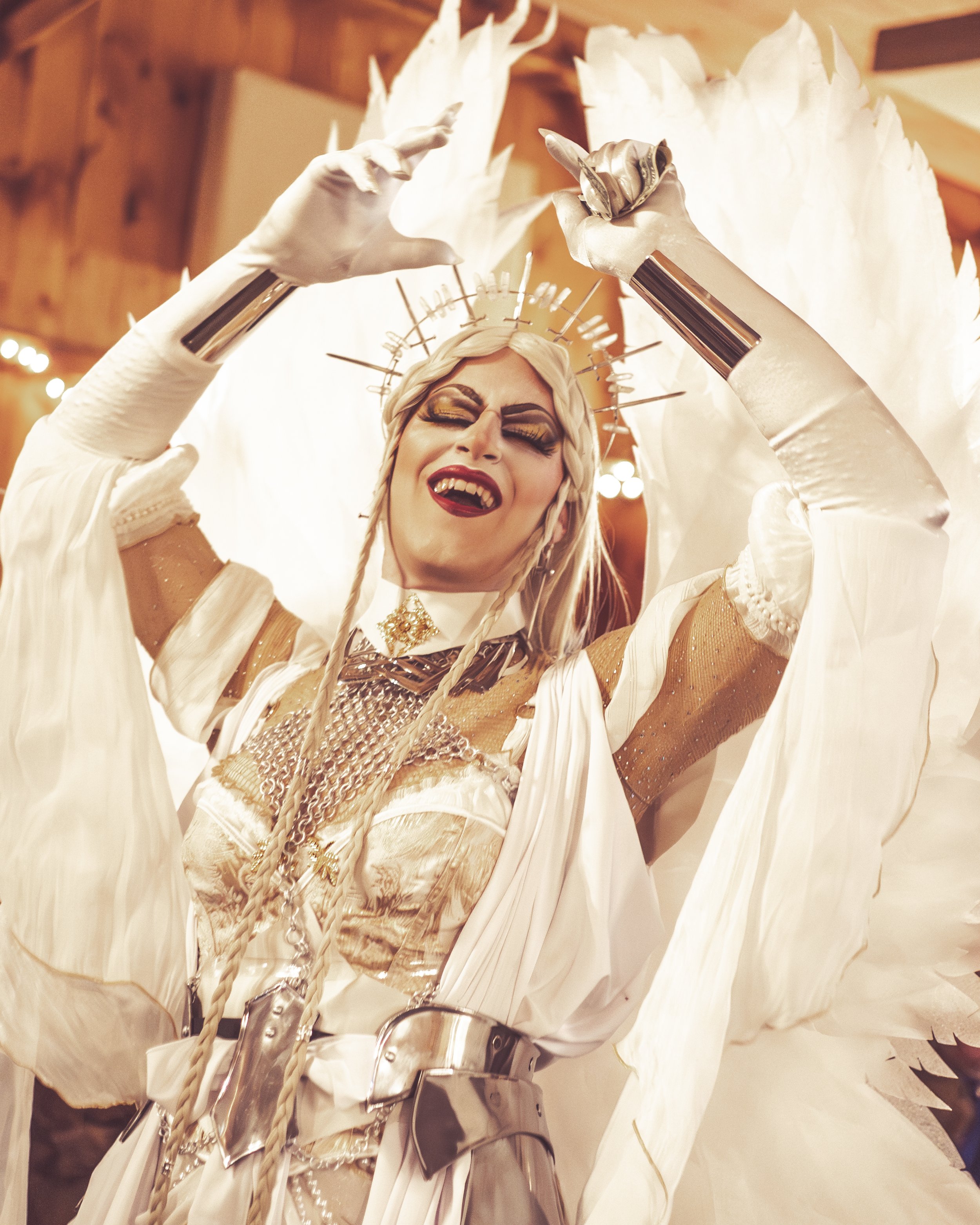 Performer dressed as an angel with large white wings, metal crown with spikes, and elaborate makeup, singing passionately with eyes closed and arms raised.