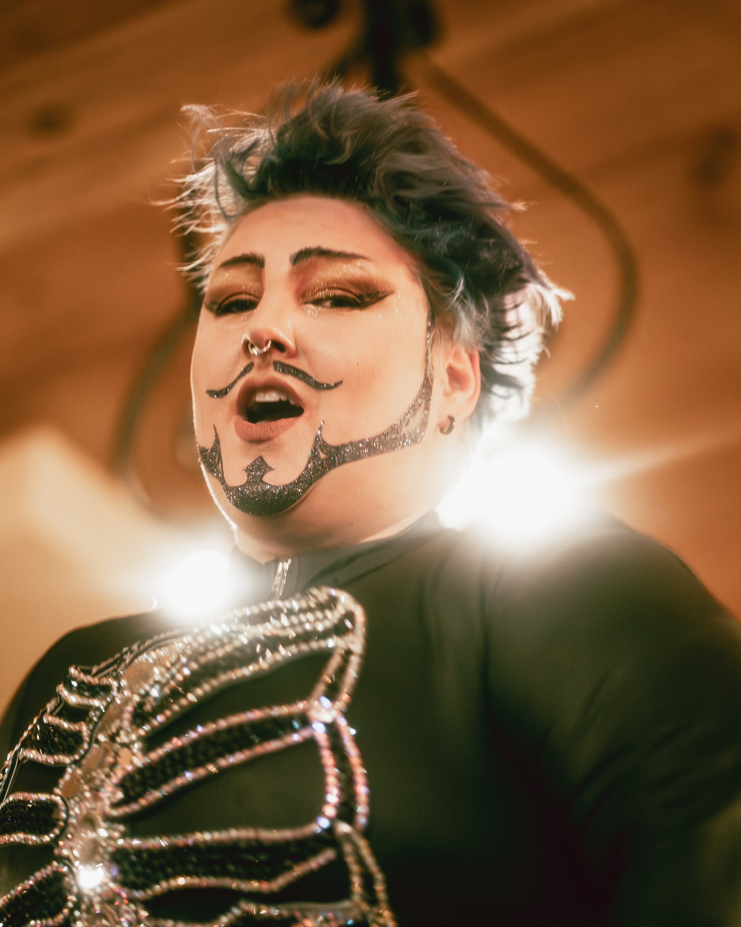 A person with short, spiky hair and bold makeup, including glittery eye shadow and face paint resembling a mustache and beard design, looking down at the camera with an expressive face, wearing black clothing with layered chains, and standing in fron