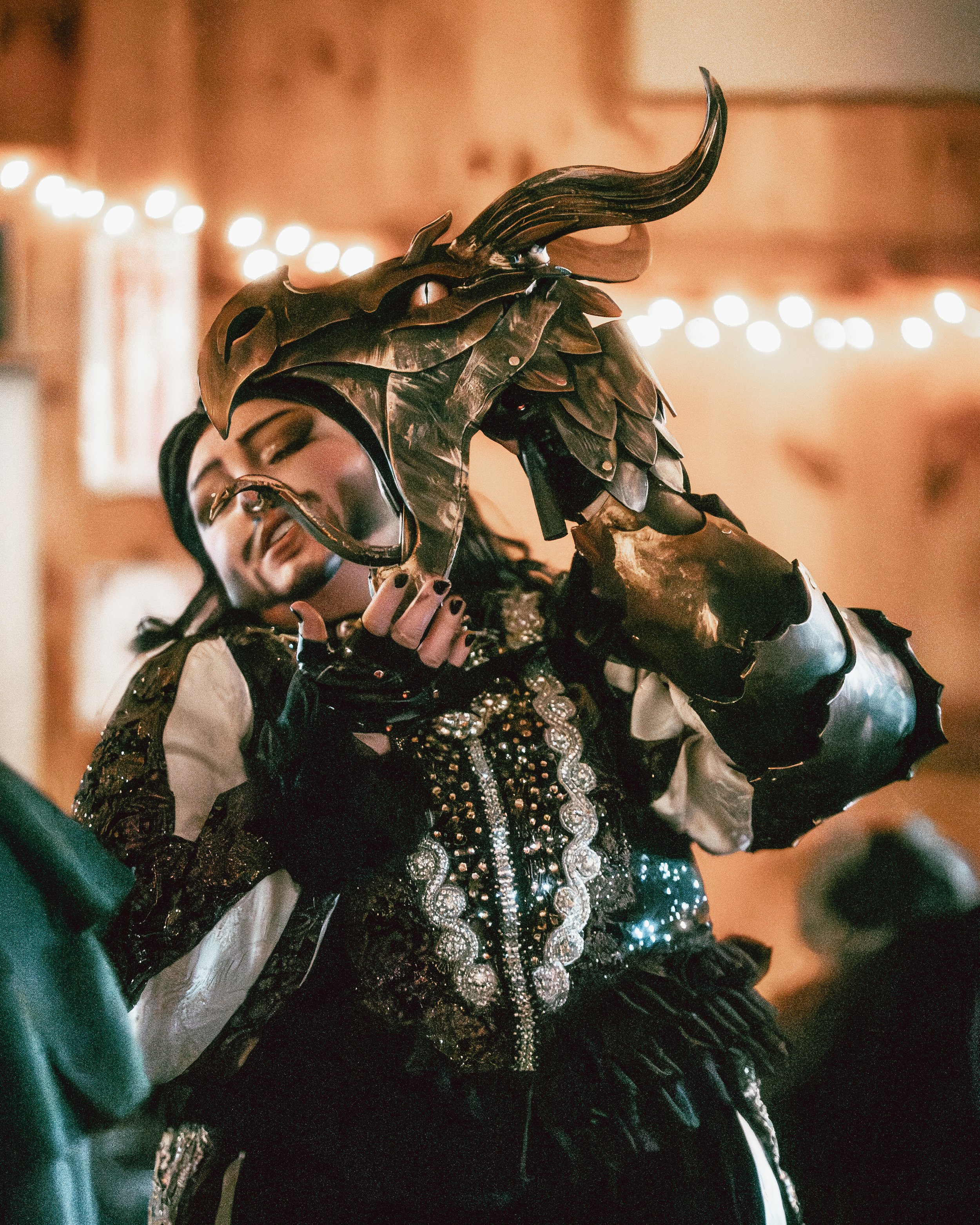 Person in a black and white costume with gold accents, wearing a dragon or lizard mask, holding a dragon or lizard mask in front of their face, in a room with warm lighting and string lights.