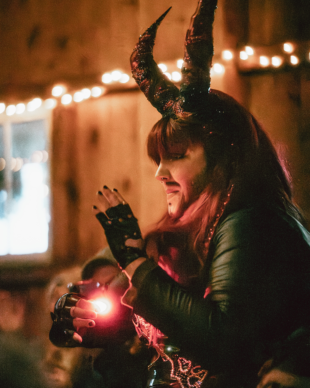 Person dressed as a demon with black and red horns, wearing black leather gloves and clothing, holding a small flashlight, in a dimly lit room decorated with string lights.