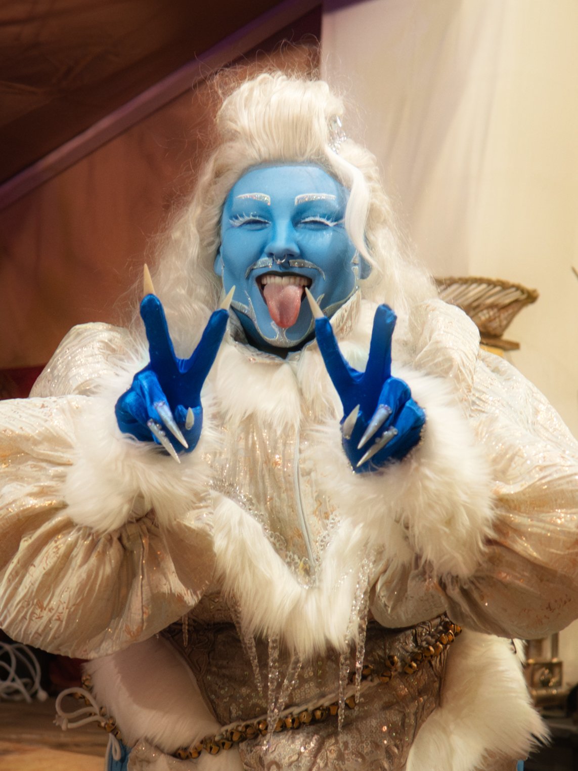 Person with blue face paint and white wig, sticking out tongue, making peace signs, in elaborate costume with furry details.