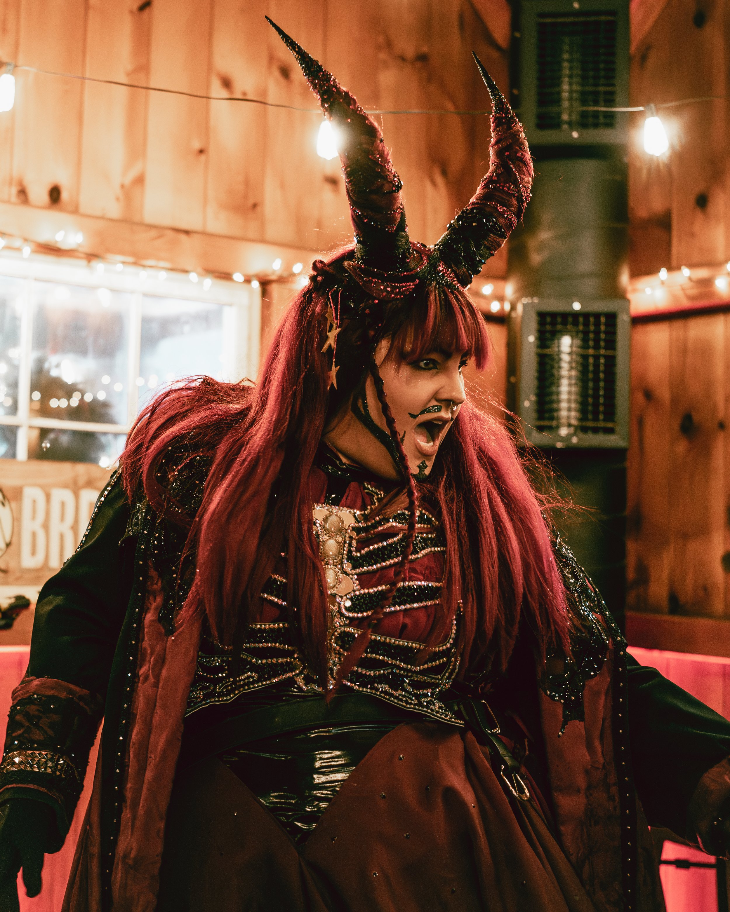 A woman dressed in a fantasy costume with large, curved, black and red horned headpiece, red hair, and makeup, inside a warmly lit wooden room with string lights.