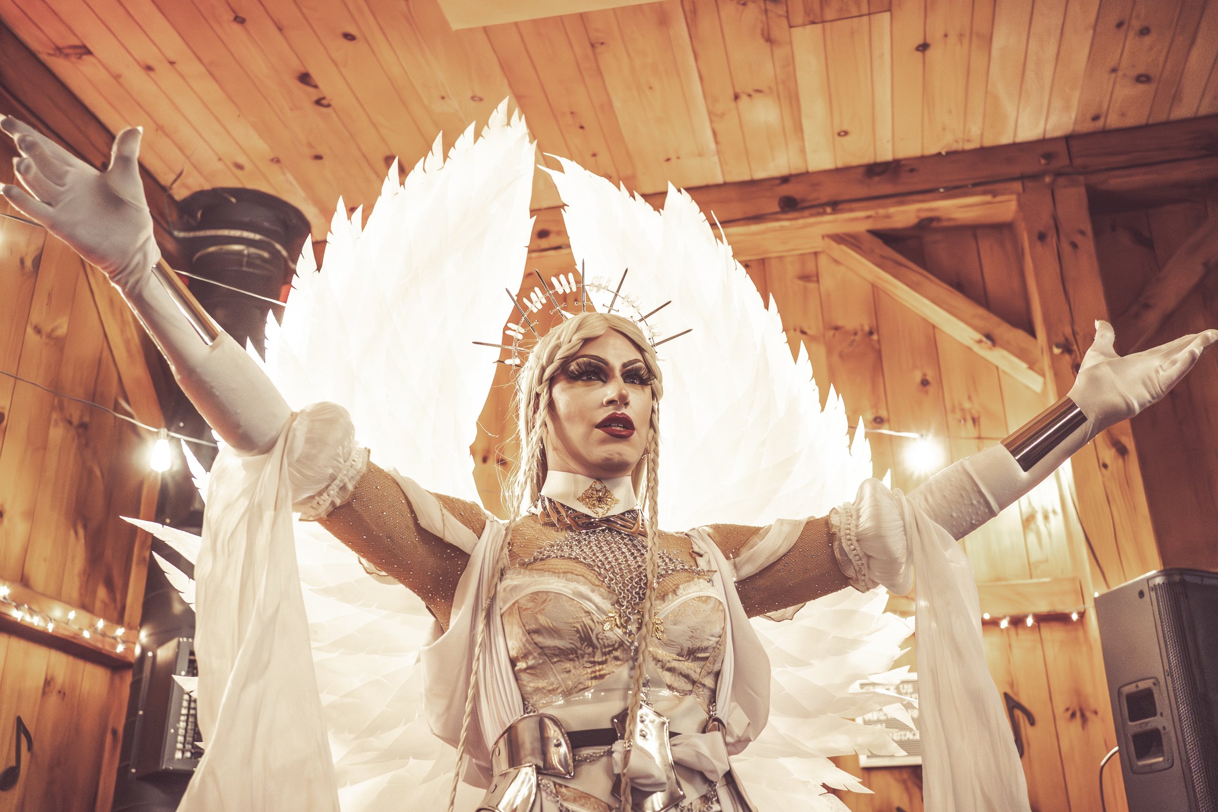A drag queen with platinum blonde hair and dark makeup performing in a wooden room with a fiery angel costume including large white feathered wings, metallic accessories, and a halo.