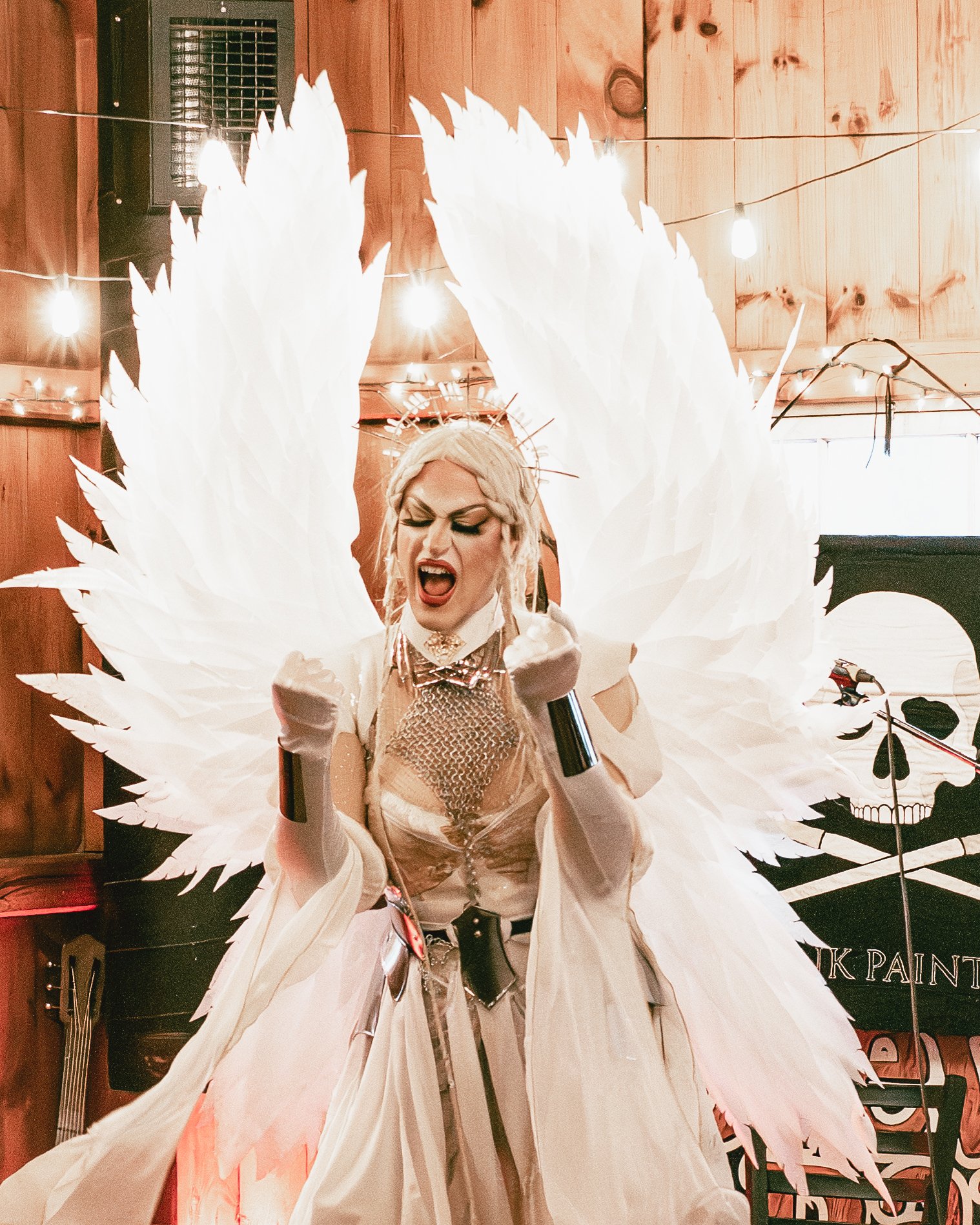 A person dressed in an elaborate costume with large white feathered wings, a braided platinum blonde wig, makeup, and holding a microphone, appears to be performing or posing in a setting with wooden walls and string lights.