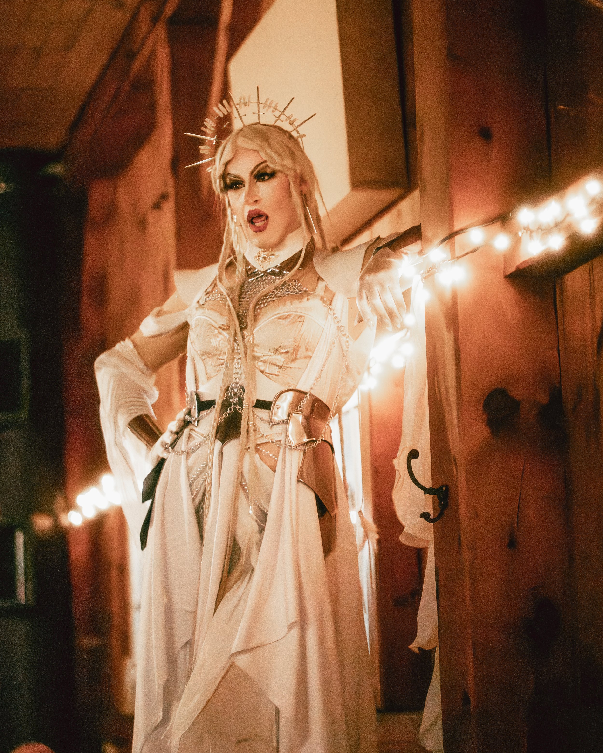 A drag queen with platinum blonde hair styled in loose waves, wearing a crown of spikes, dramatic makeup, and an elaborate beige costume with chains and metallic embellishments. She is posing with an intense expression, inside a wooden room with stri