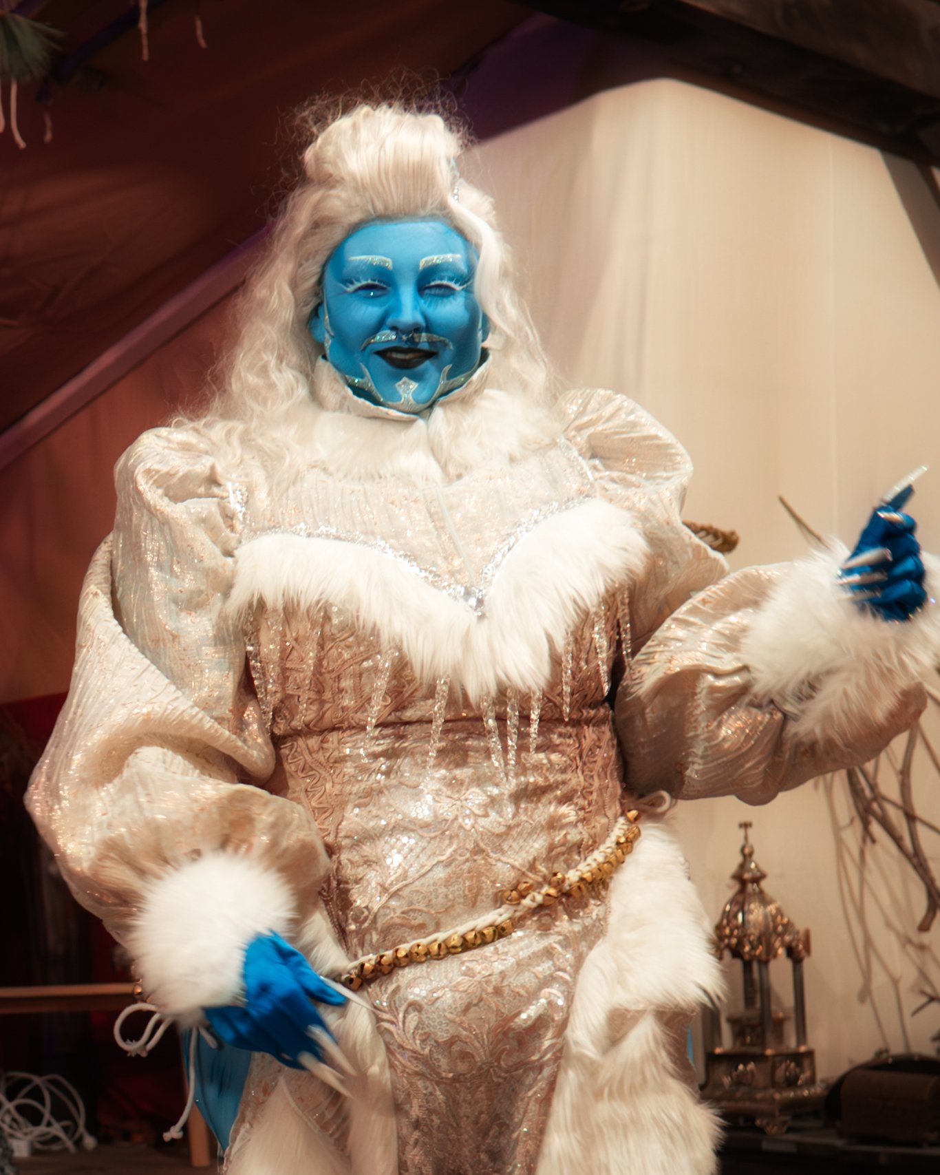 Person dressed in an elaborate costume with blue face paint, white costume with fur trim, and long white hair, smiling and holding a cigarette.