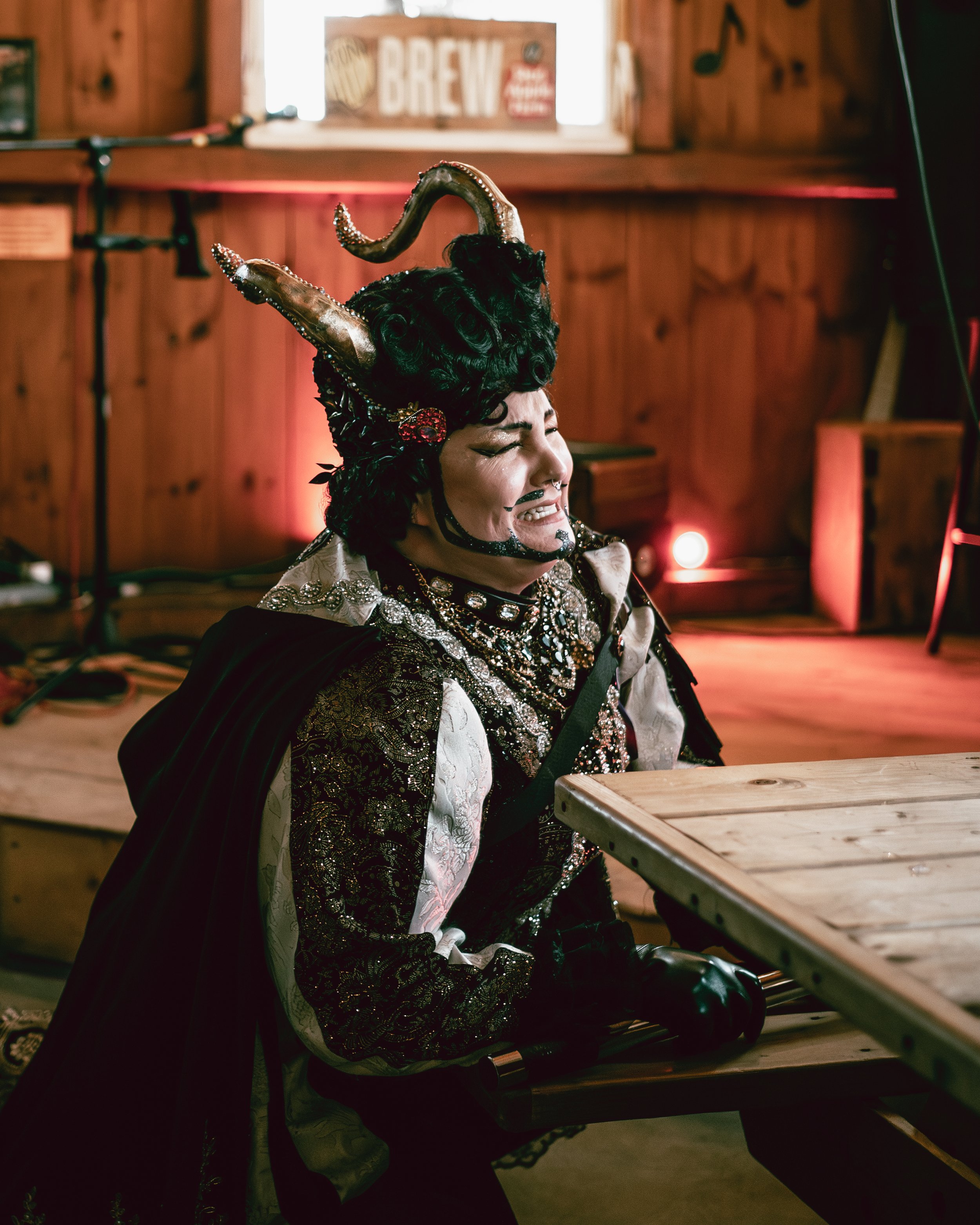 Person in elaborate costume with horns, makeup, and jewelry, sitting at a wooden table in a dimly lit, rustic room.