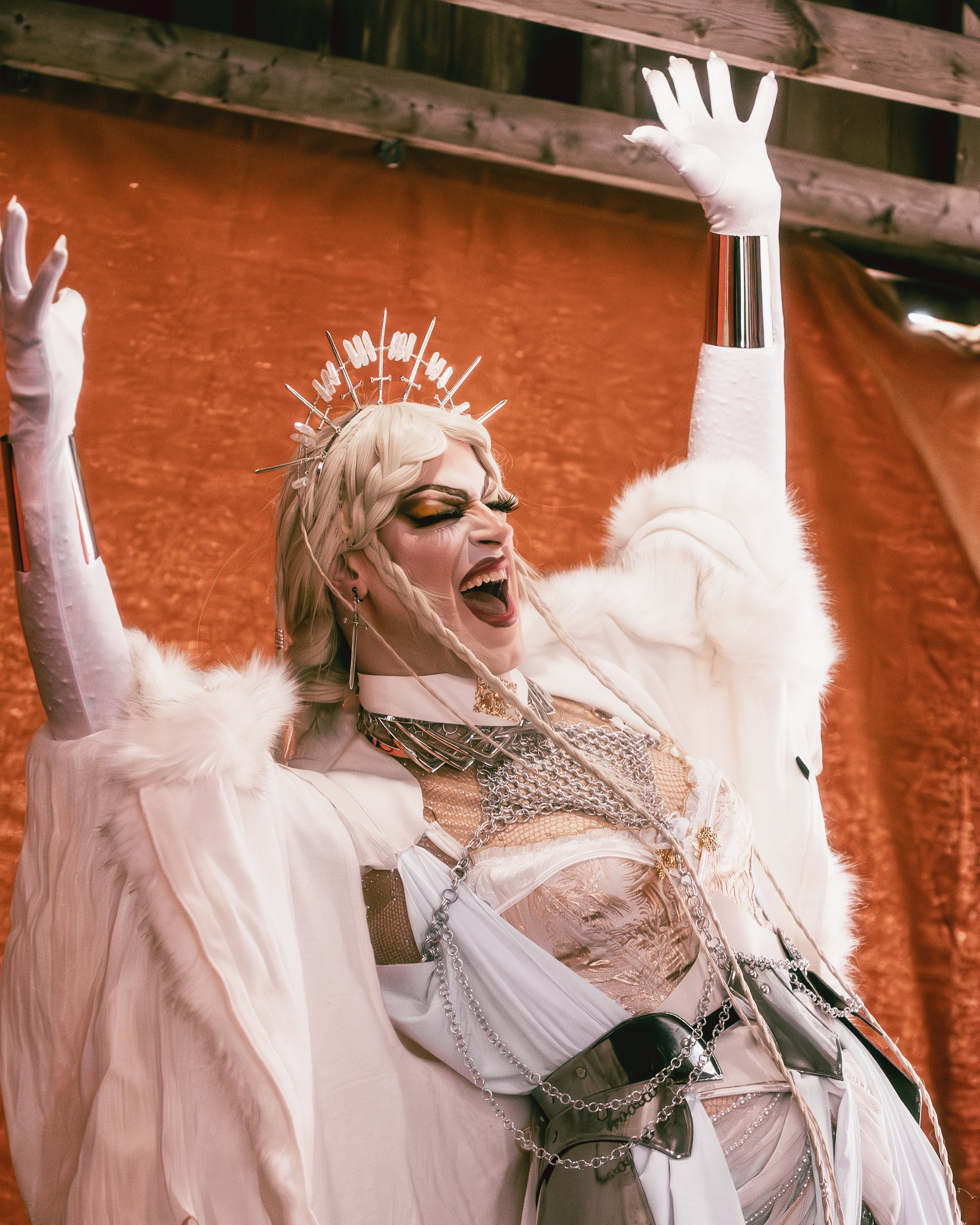 Performer dressed in elaborate costume with chains, makeup, and headpiece, with arms raised and mouth open, against a reddish background.