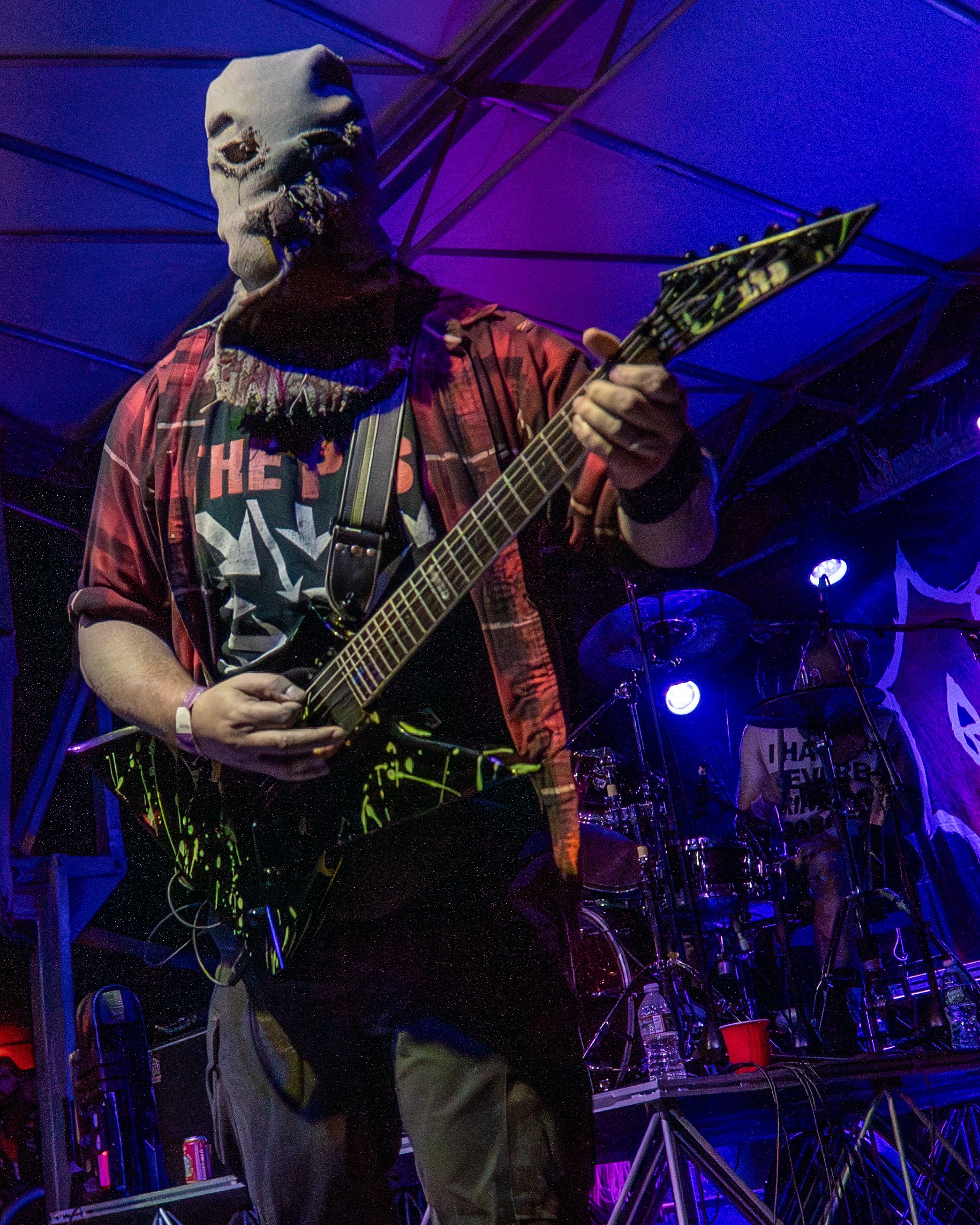 A musician wearing a creature mask playing an electric guitar on stage with drums in the background, illuminated by purple and blue stage lighting.