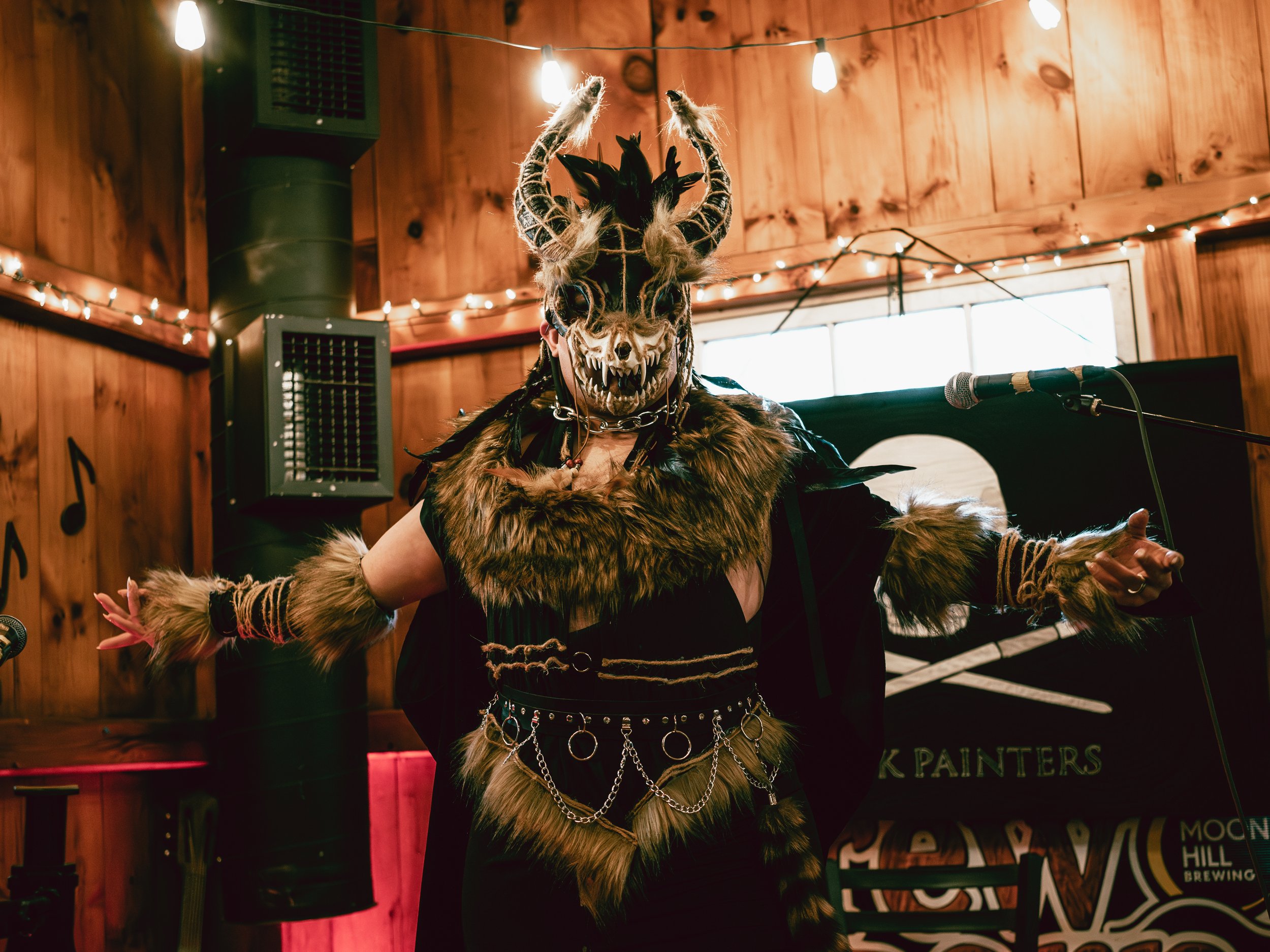 Person in elaborate monster costume with horns and fur, performing on stage with a microphone in a wood-paneled room decorated with string lights.