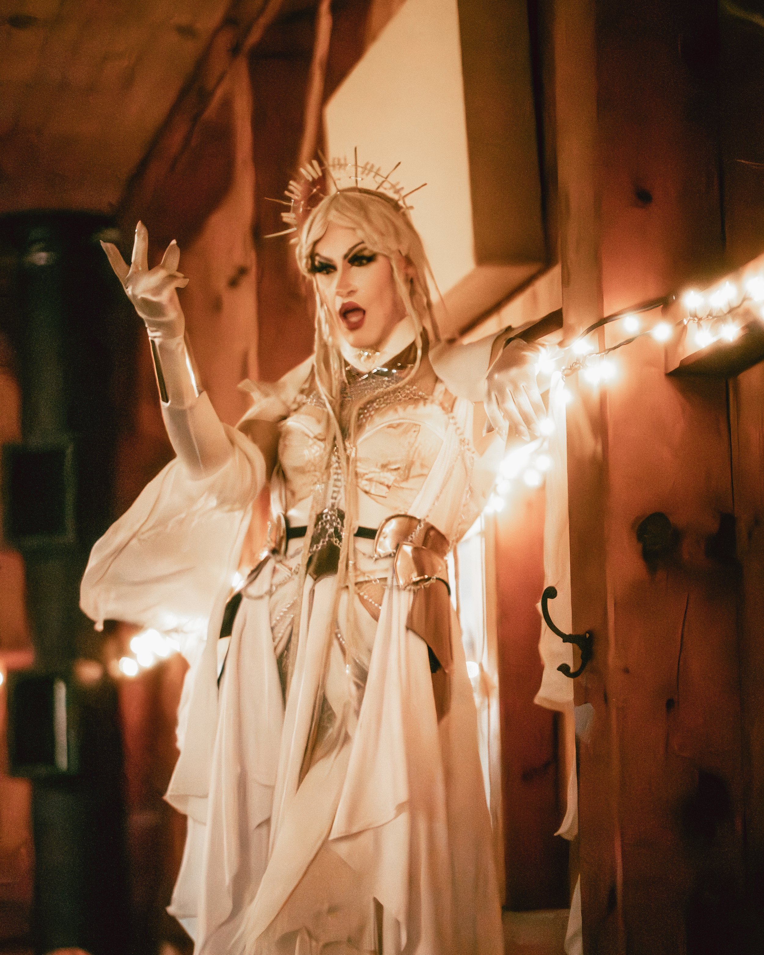 A drag performer in a cream-colored outfit with elaborate makeup, a crown, and long gloves, standing on a wooden staircase with string lights, striking a dramatic pose.