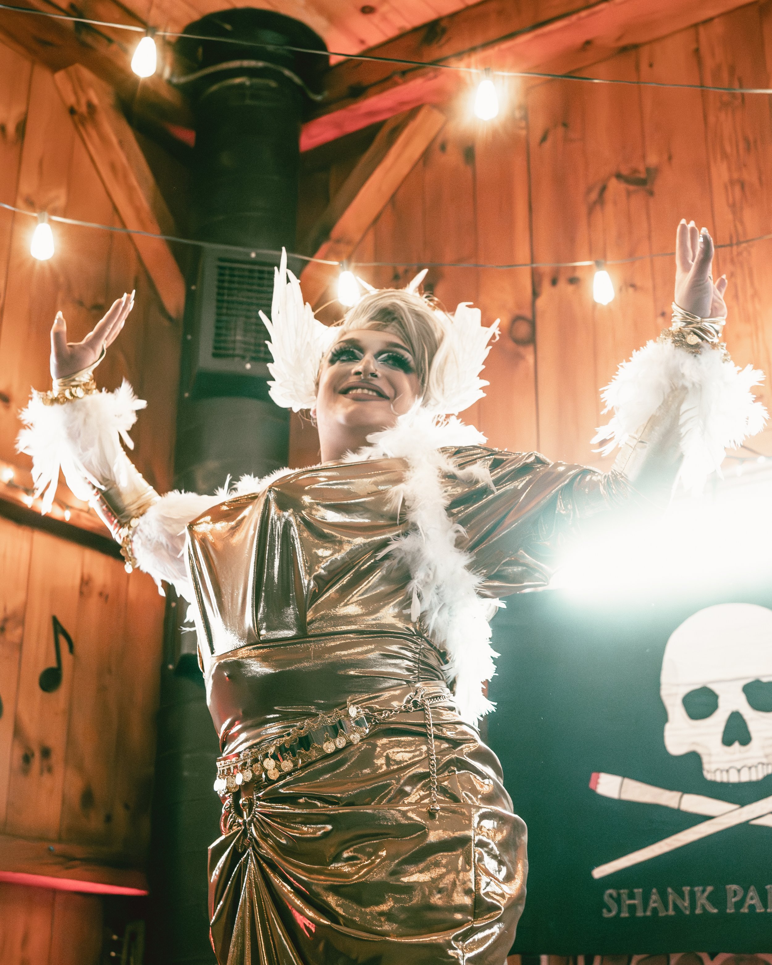 A person dressed in a shiny, gold outfit with white feathered accessories, wearing a large feathered headpiece, is performing or posing in front of a wooden wall with string lights overhead and a black banner with a skull and crossbones design.