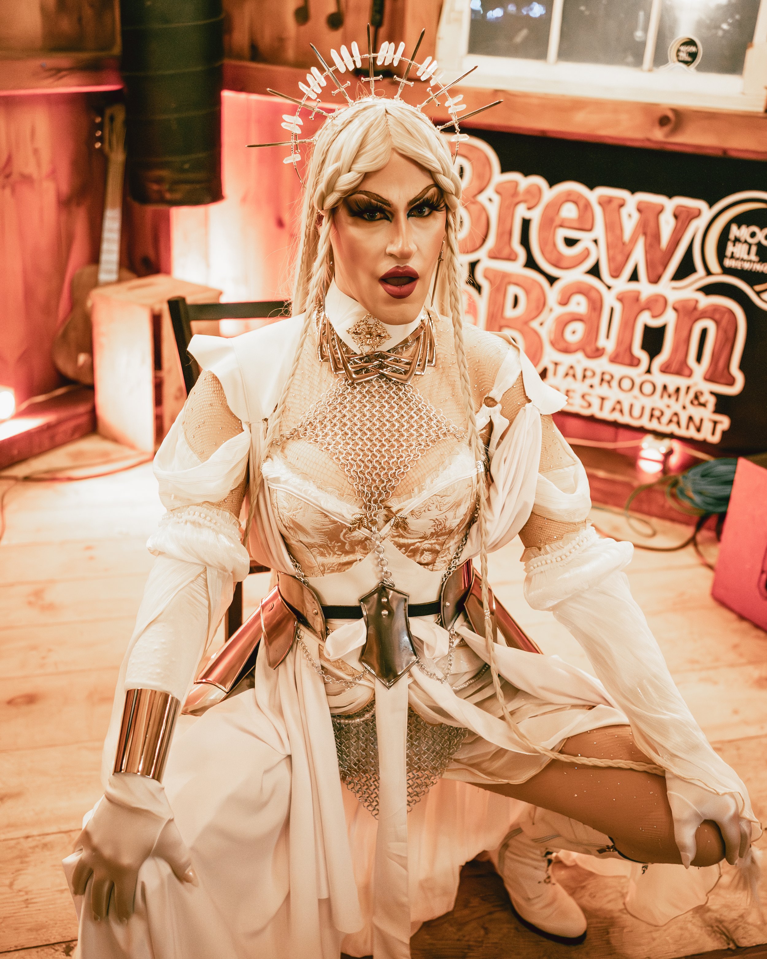 Person in elaborate costume with metallic accessories and a crown, in a wooden room with a sign for a brew barn in the background.